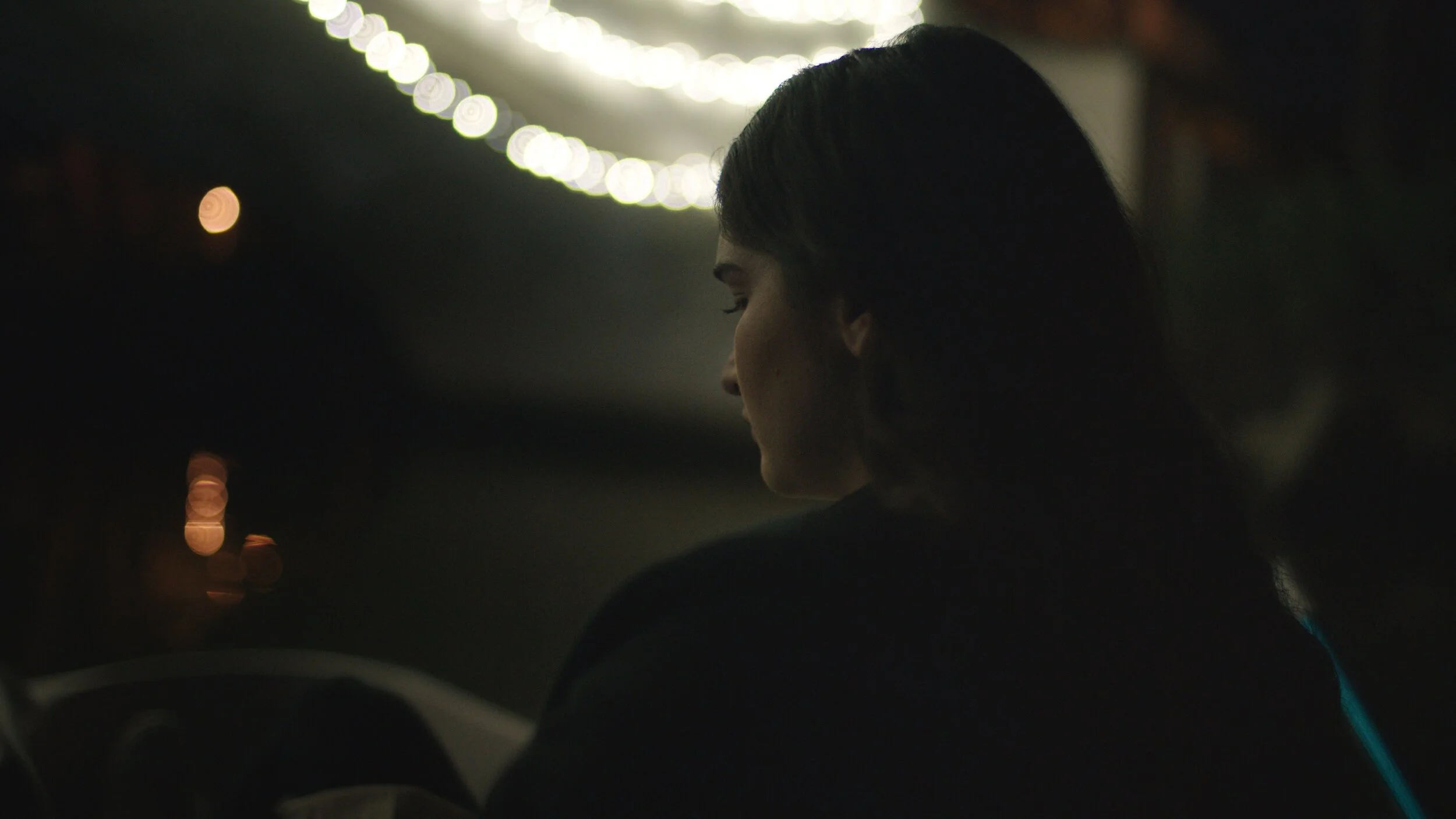 Still from the short film, FOR GOOD. At night, a woman with dark hair sits outside. Her profile is illuminated by the soft glow of string lights.