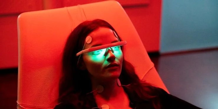 Still from the short film, THE ONE. In a room with red and blue lighting, a woman (Madeline Brewer) reclines in a chair with a medical device on her forehead emitting greenish-white light.