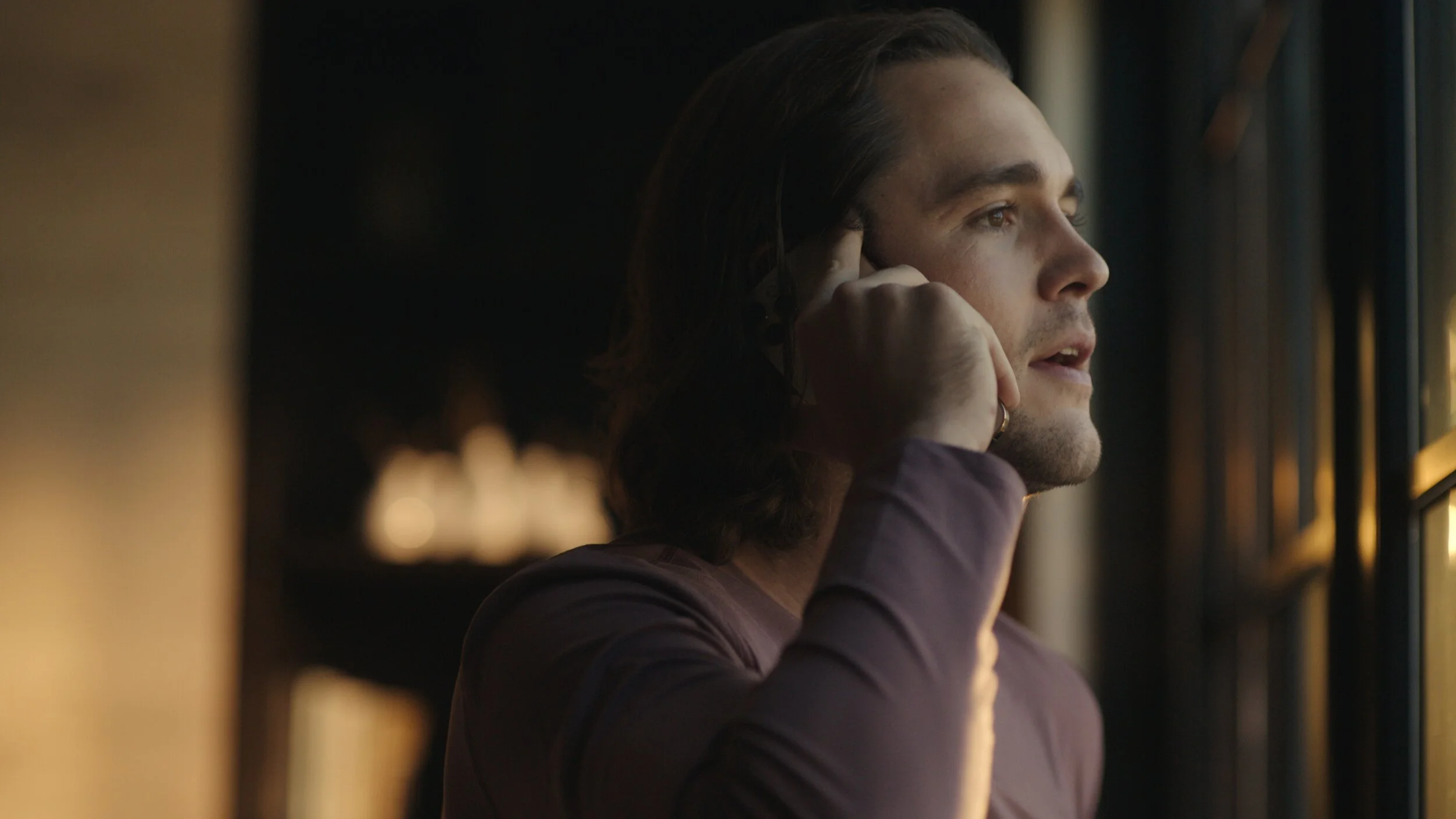 Still from the short film, FOR GOOD. A man with long hair looks out a window during sunset, holding a phone to his ear.