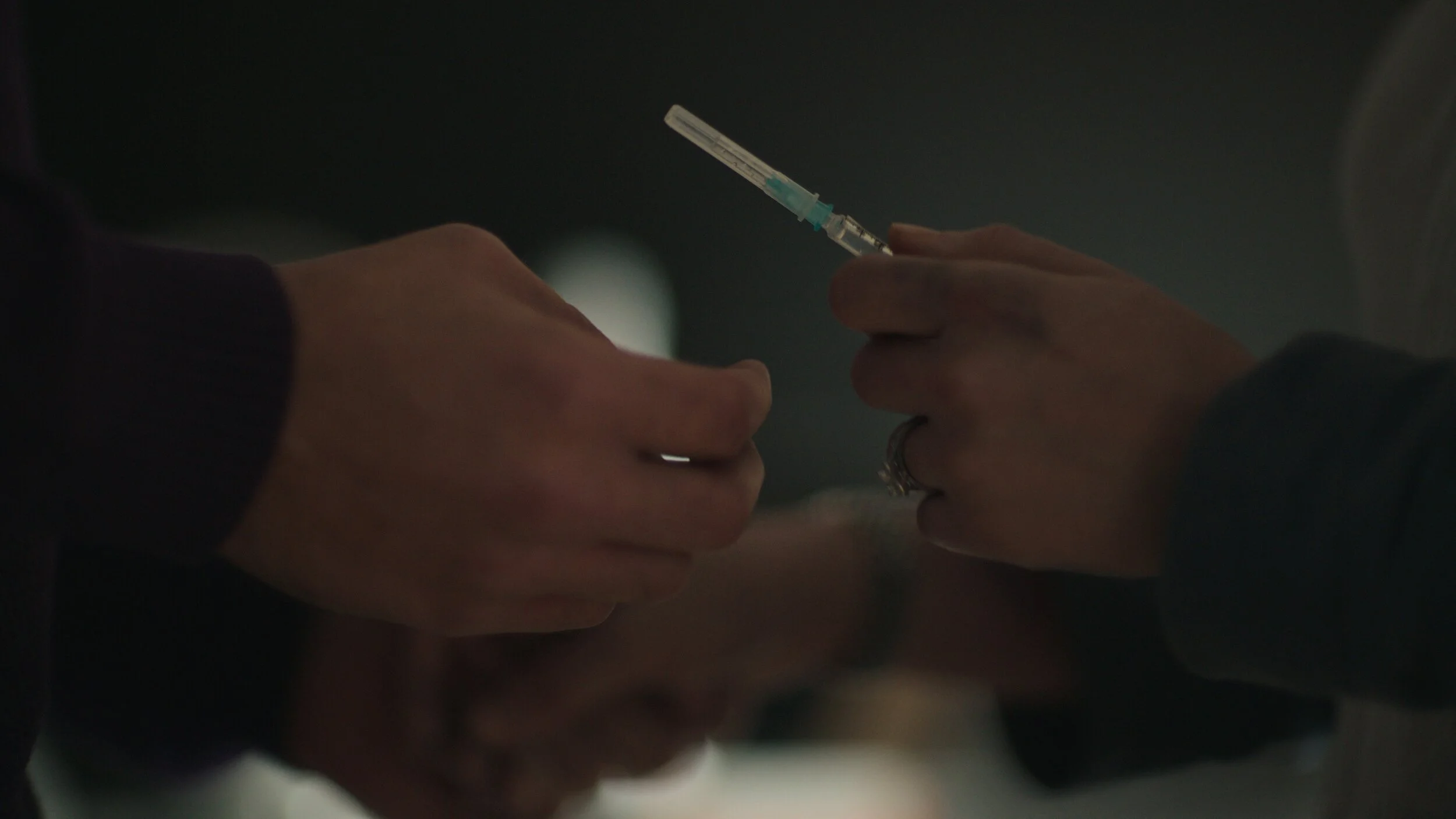 Still from the short film, FOR GOOD. Close-up of hands exchanging a syringe, with one person preparing to administer an injection.