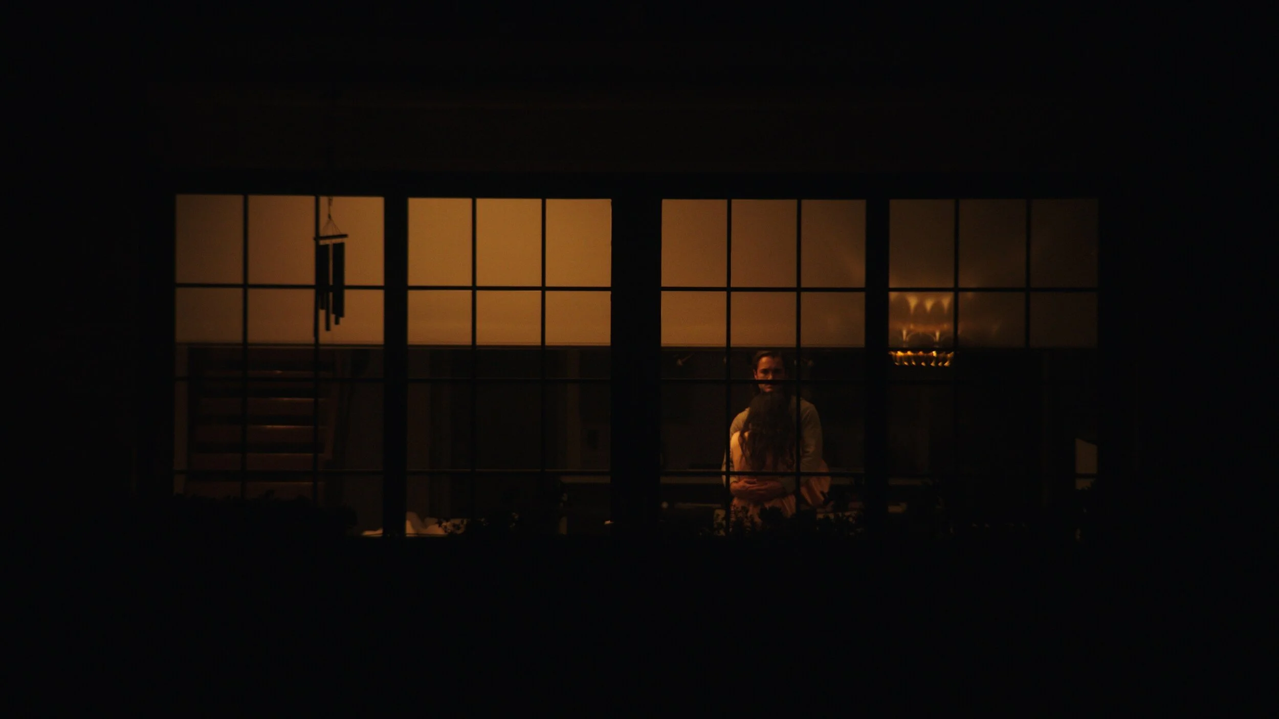 Still from the short film, FOR GOOD. A person is standing inside a dimly lit room, seen through a large window with multiple panes. The room has a warm, orange glow from interior lighting.