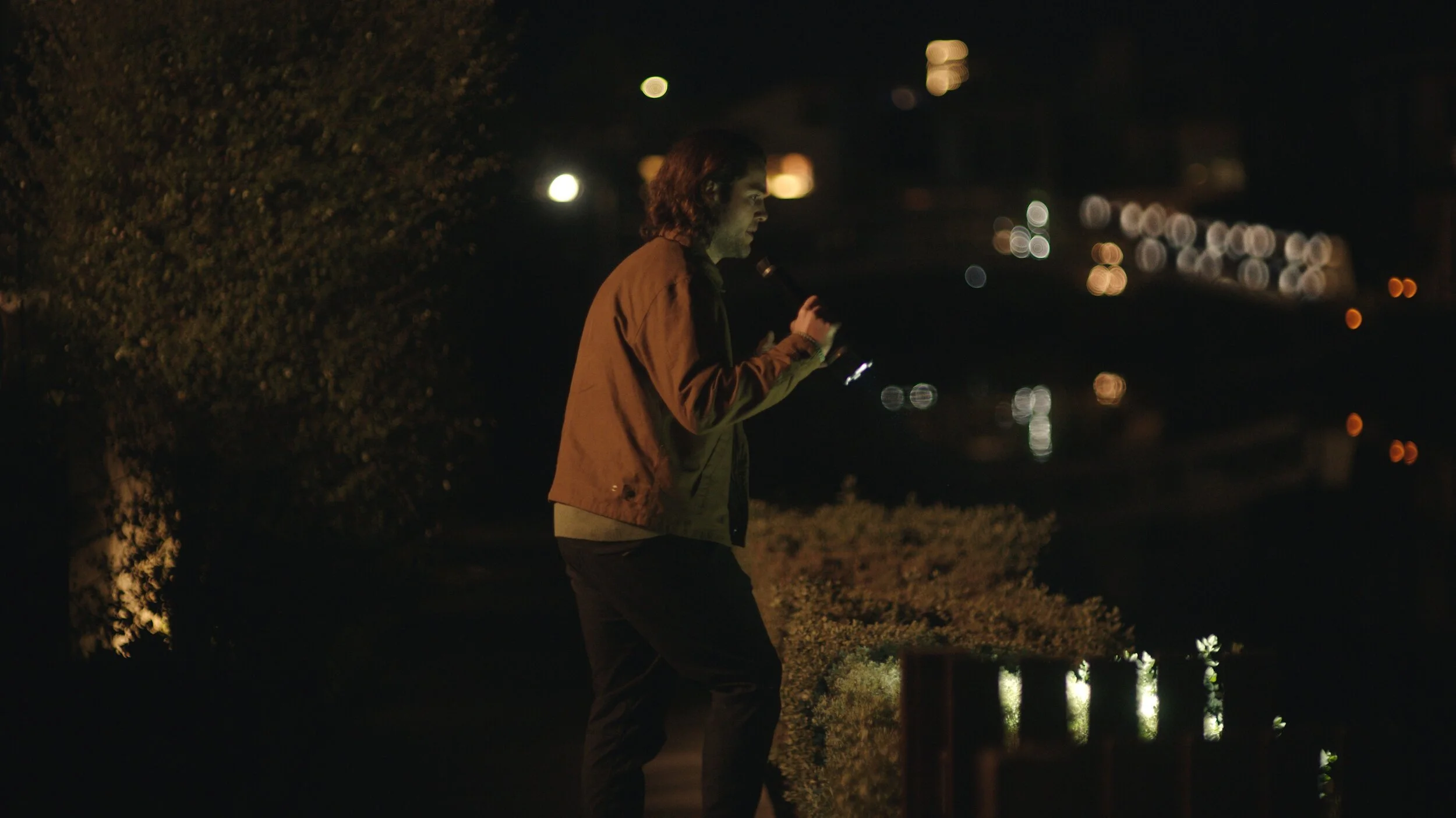 Still from the short film, FOR GOOD. At night, a man with dark hair stands alone, holding a phone. He wears a brown jacket and dark pants. The background is blurred with various city lights reflecting across the water.
