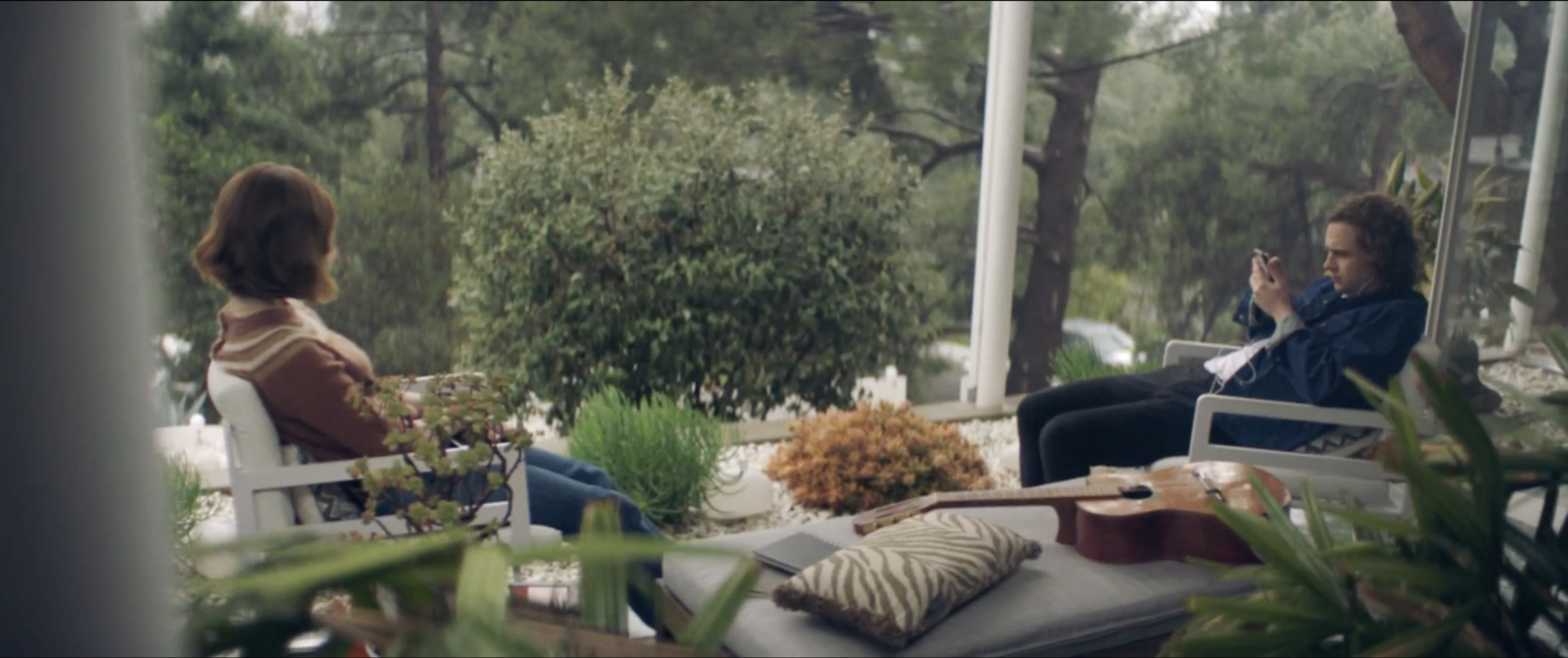 Still from the short film, SILVERTONE. A woman (Kerris Dorsey) and a man (Logan Miller) sit on a porch surrounded by plants and trees. The woman is facing away, and the man is looking at his phone. An acoustic guitar rests on a table nearby.