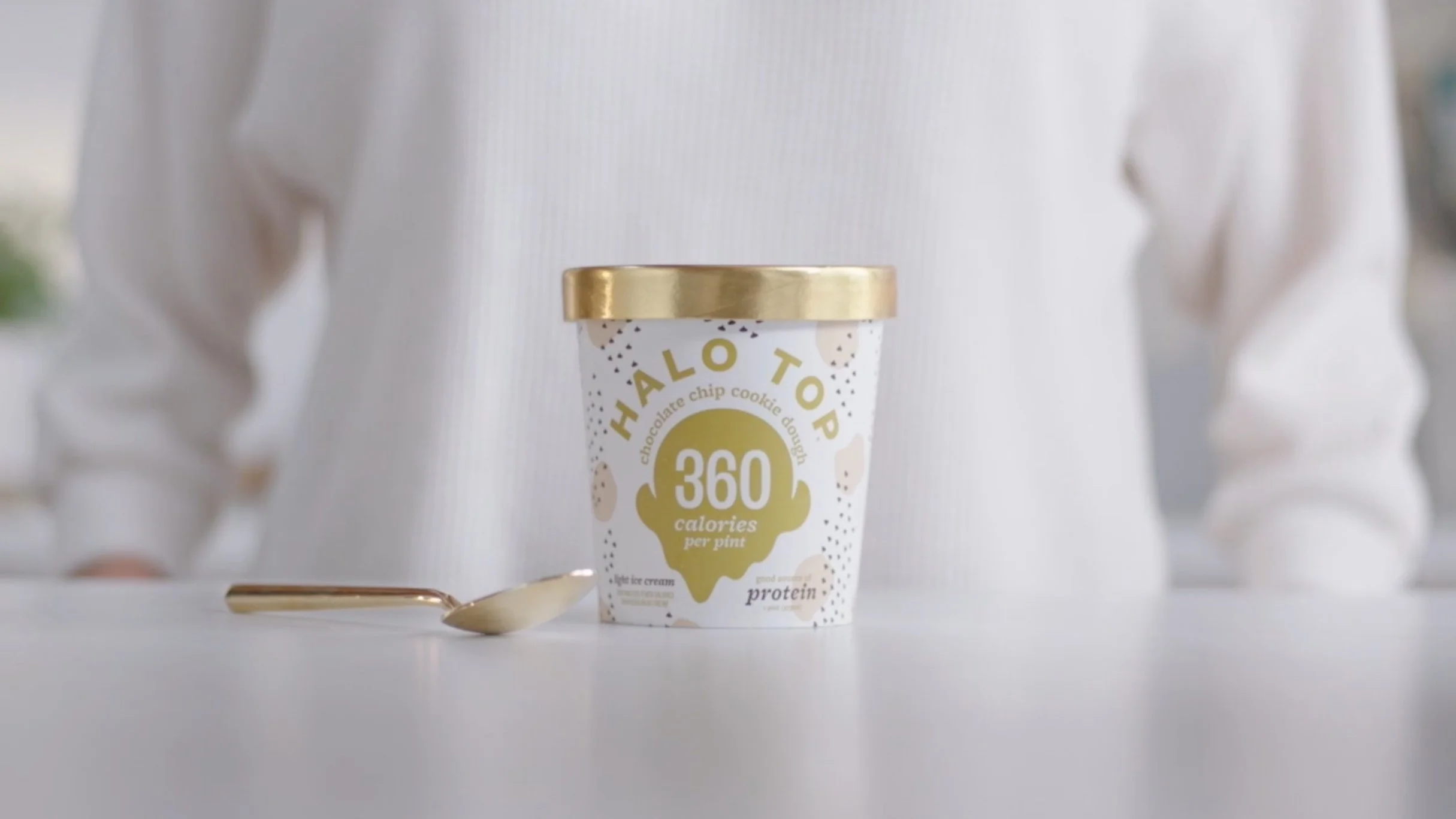 A pint of Halo Top chocolate chip cookie dough ice cream with a gold lid rests on a white surface near a spoon.