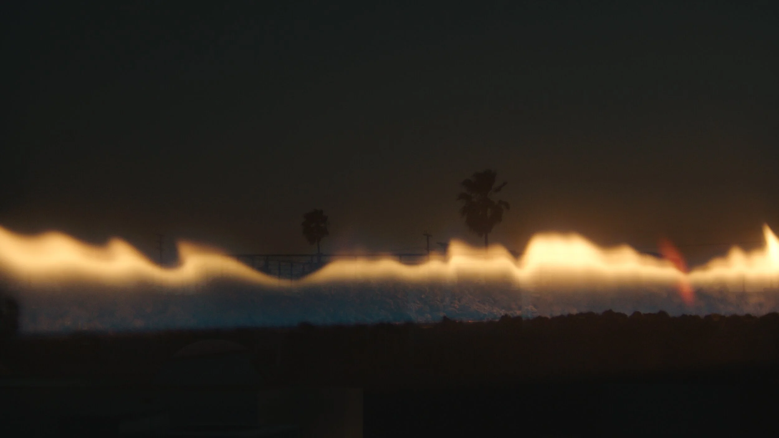 Still from the short film, FOR GOOD. Tall flames from an outdoor fire pit illuminate the silhouettes of palm trees against the night sky.