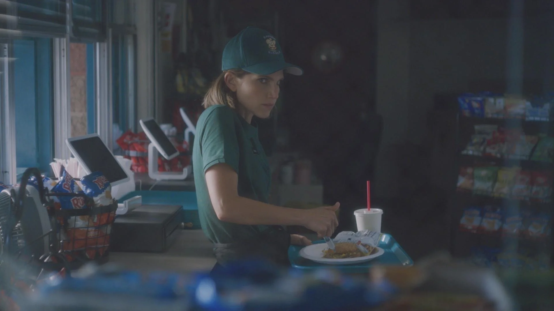 Still from the short film, SUSIE SEARCHES. A young woman (Sophie Kargman) wearing a green uniform and cap stands at a convenience store checkout counter with a food tray and a soft drink.