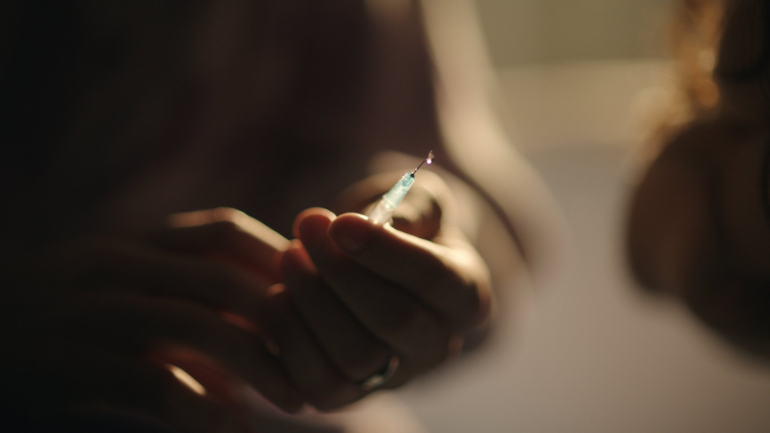 Still from the short film, FOR GOOD. Close-up of a person's hand holding a syringe with a needle, poised to inject a substance.