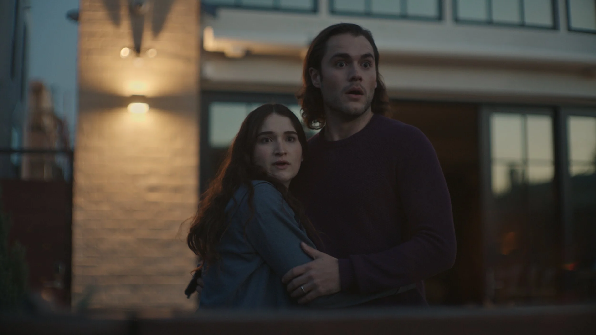 Still from the short film, FOR GOOD. A young woman and a man with long hair are standing outside a modern home at dusk, looking surprised as they embrace.