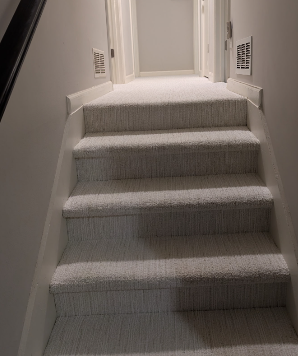 Carpeted staircase leading up to a landing with a closed door in the background, white walls, and air vents on both sides.