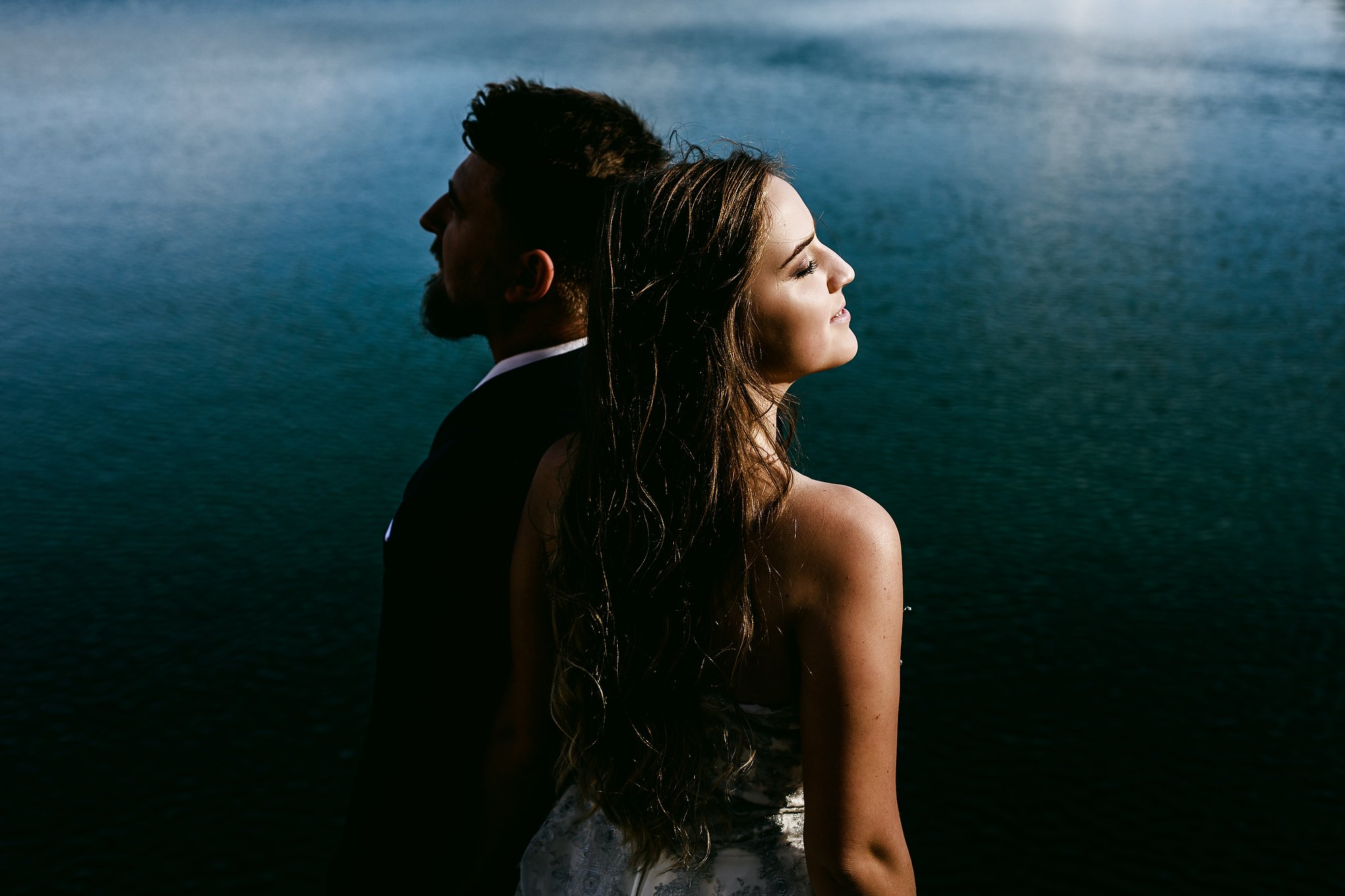 A man in a suit standing behind a woman in a dress, both looking away from the camera, with a body of water in the background.