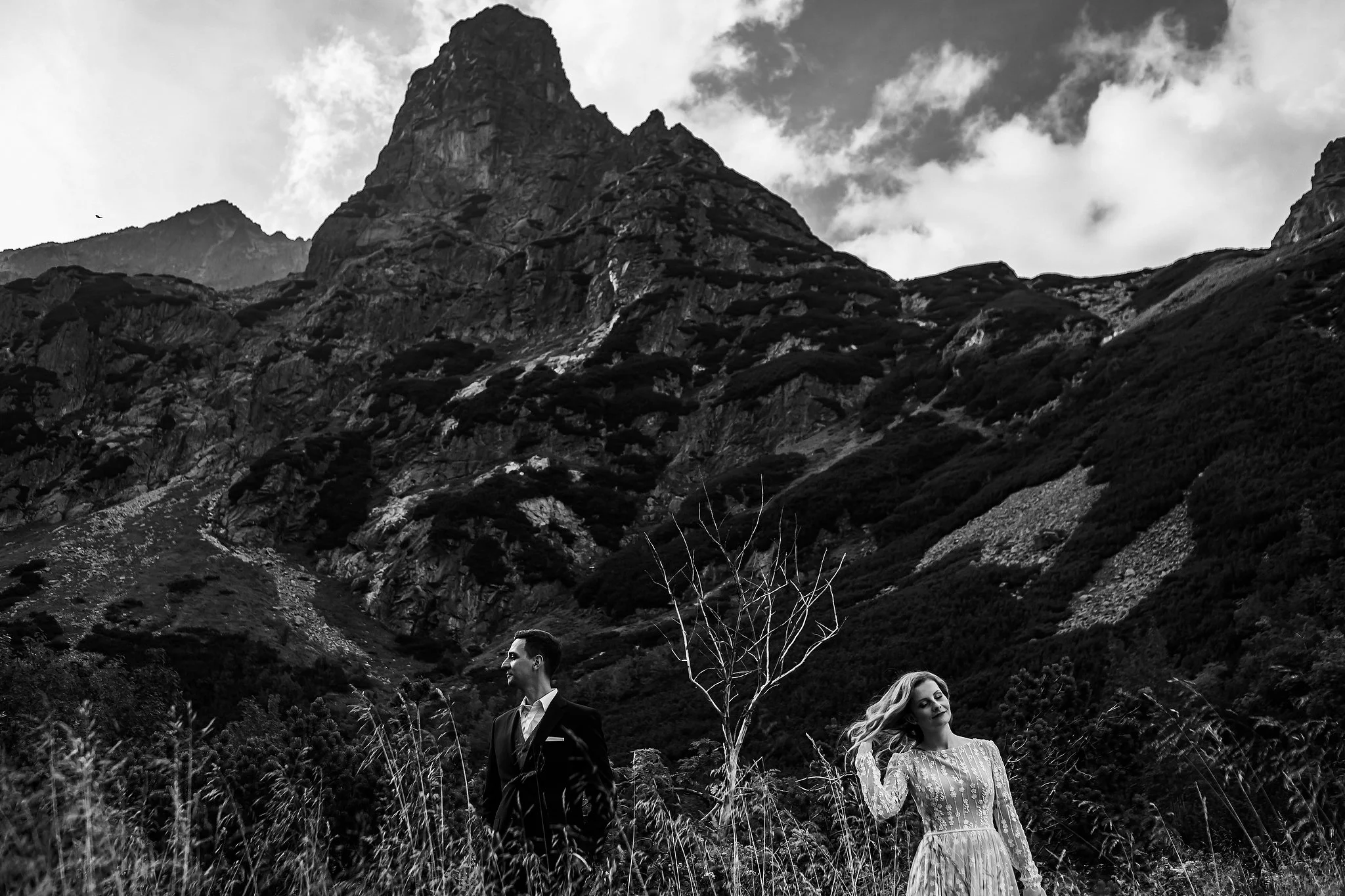 Black and white photo of a man in a suit and a woman in a dress standing in a natural landscape with tall grass and a mountain in the background.