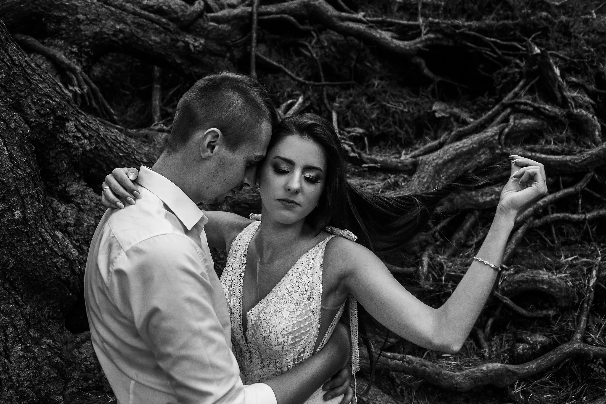 A black and white photo of a couple embracing outdoors near a large tree with exposed roots, with the woman holding her hair and the man close to her face.