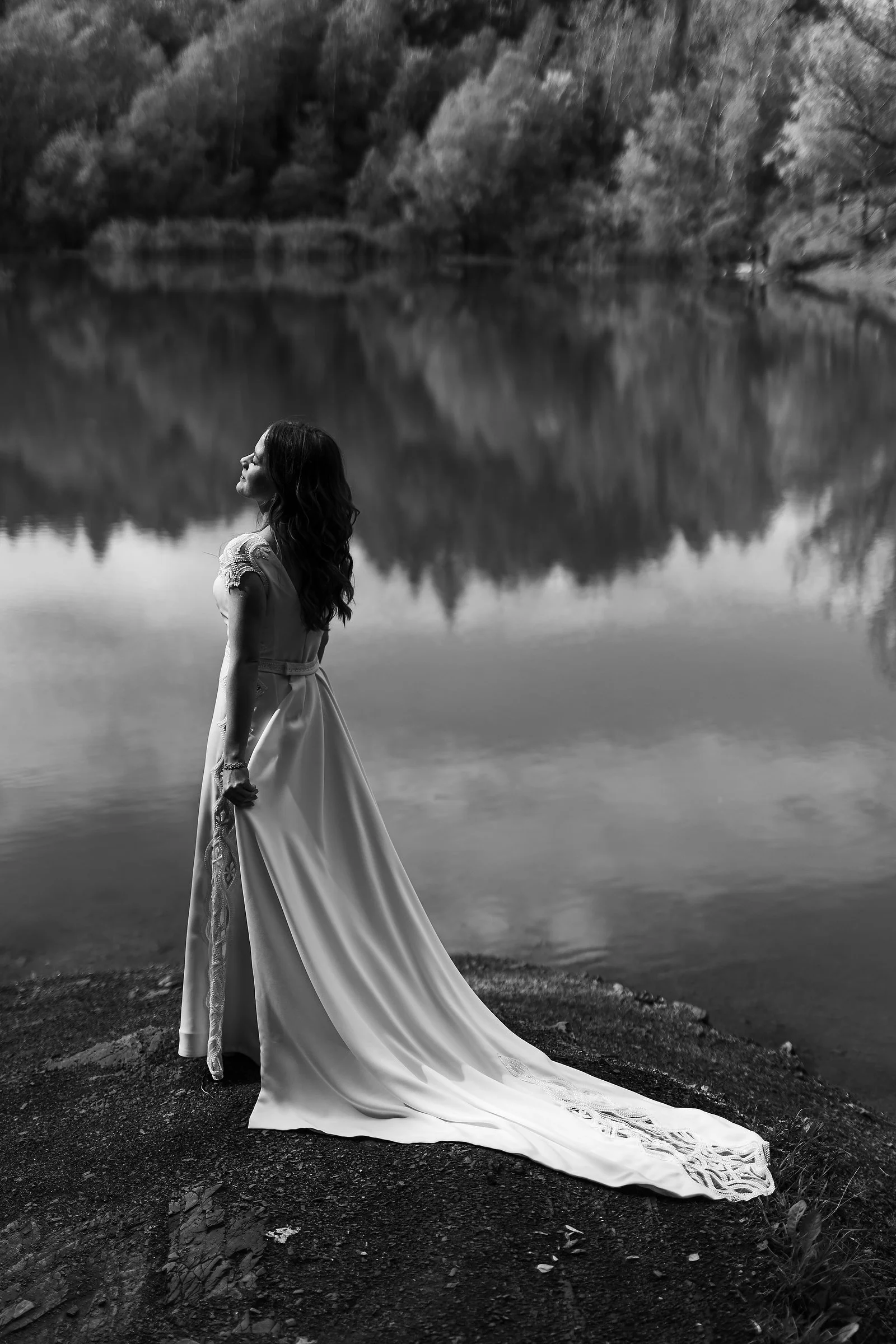 Black and white photo of a woman in a wedding dress standing by a lake, with trees and reflection in the water.