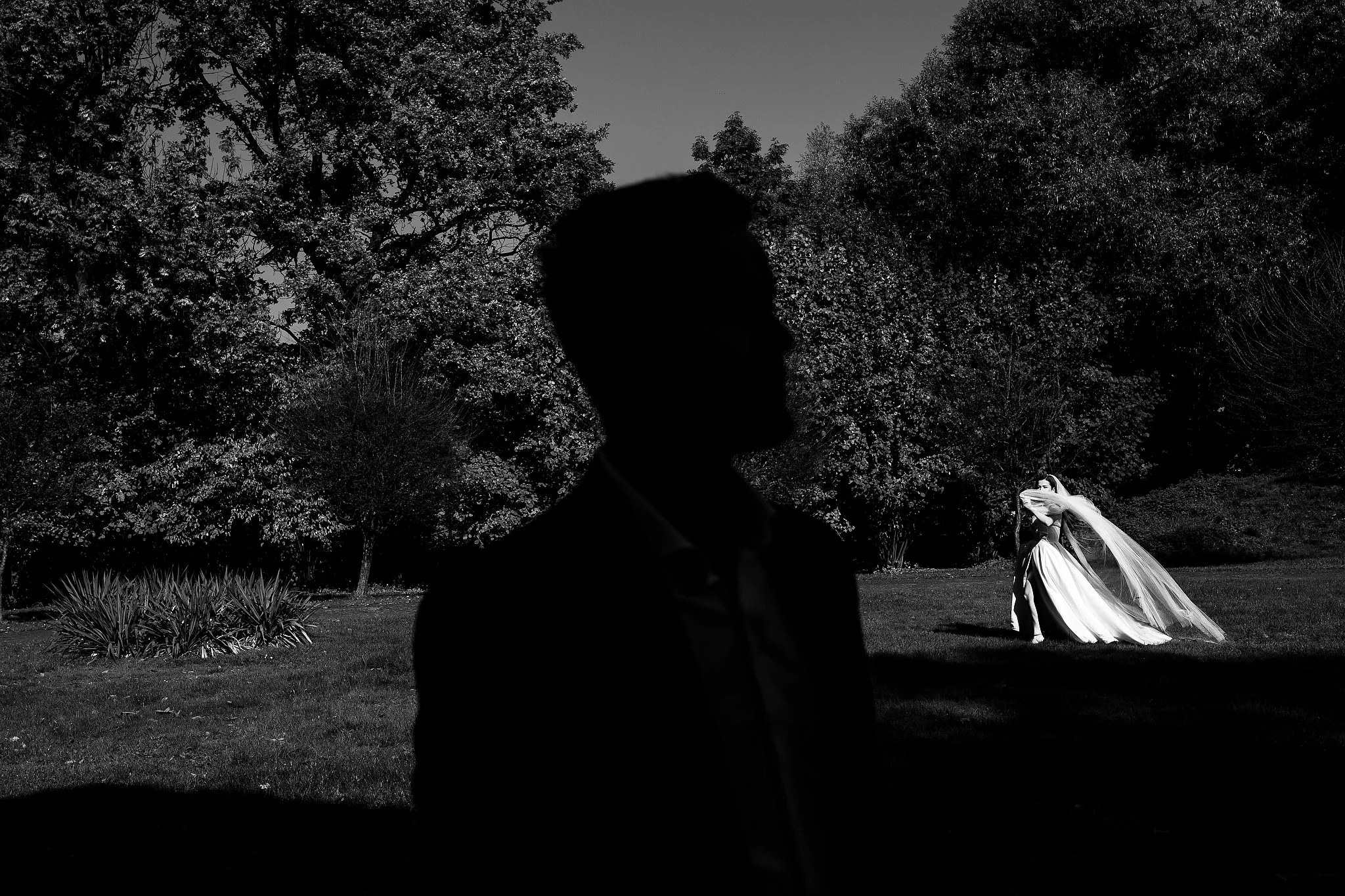 A silhouette of a man in the foreground with a wedding couple in the background, outdoors, trees and grass, black and white photo.