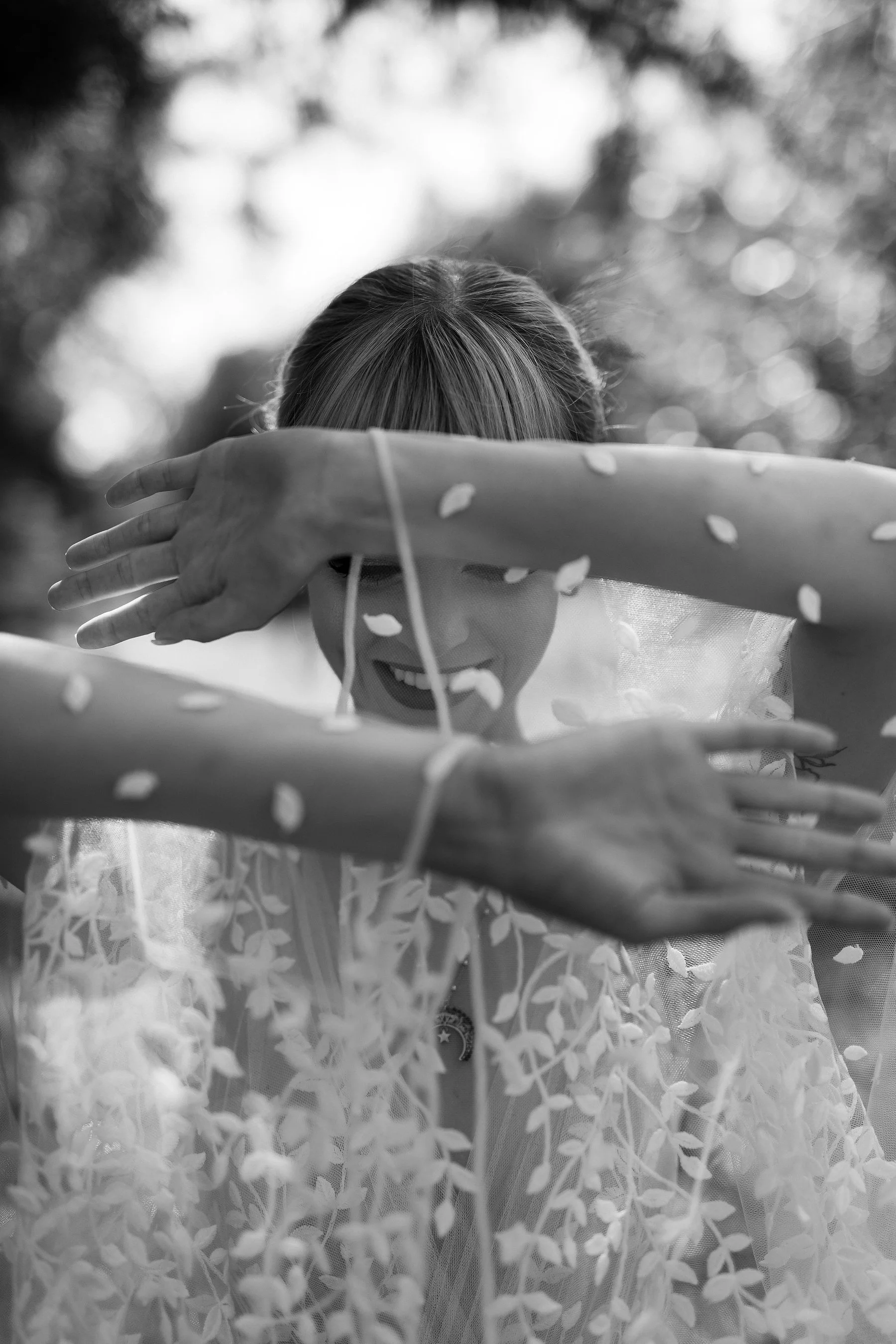 A woman in a translucent dress with a floral pattern uses her arms to create a frame around her face. She is smiling, with her eyes closed, and facing the camera. The background is blurred with trees and sky.