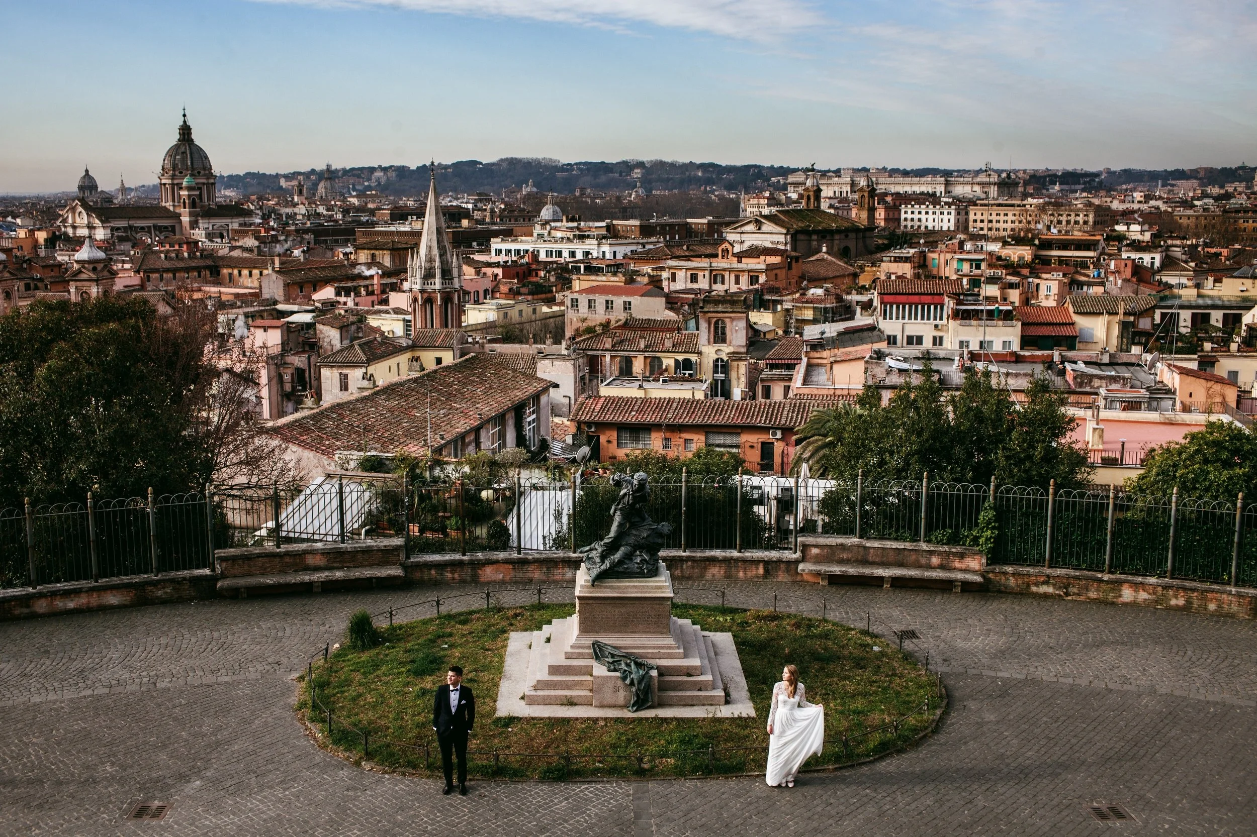 A bride in a white wedding dress and a groom in a tuxedo stand near a statue on a hilltop with a cityscape in the background, featuring domed and spired buildings.