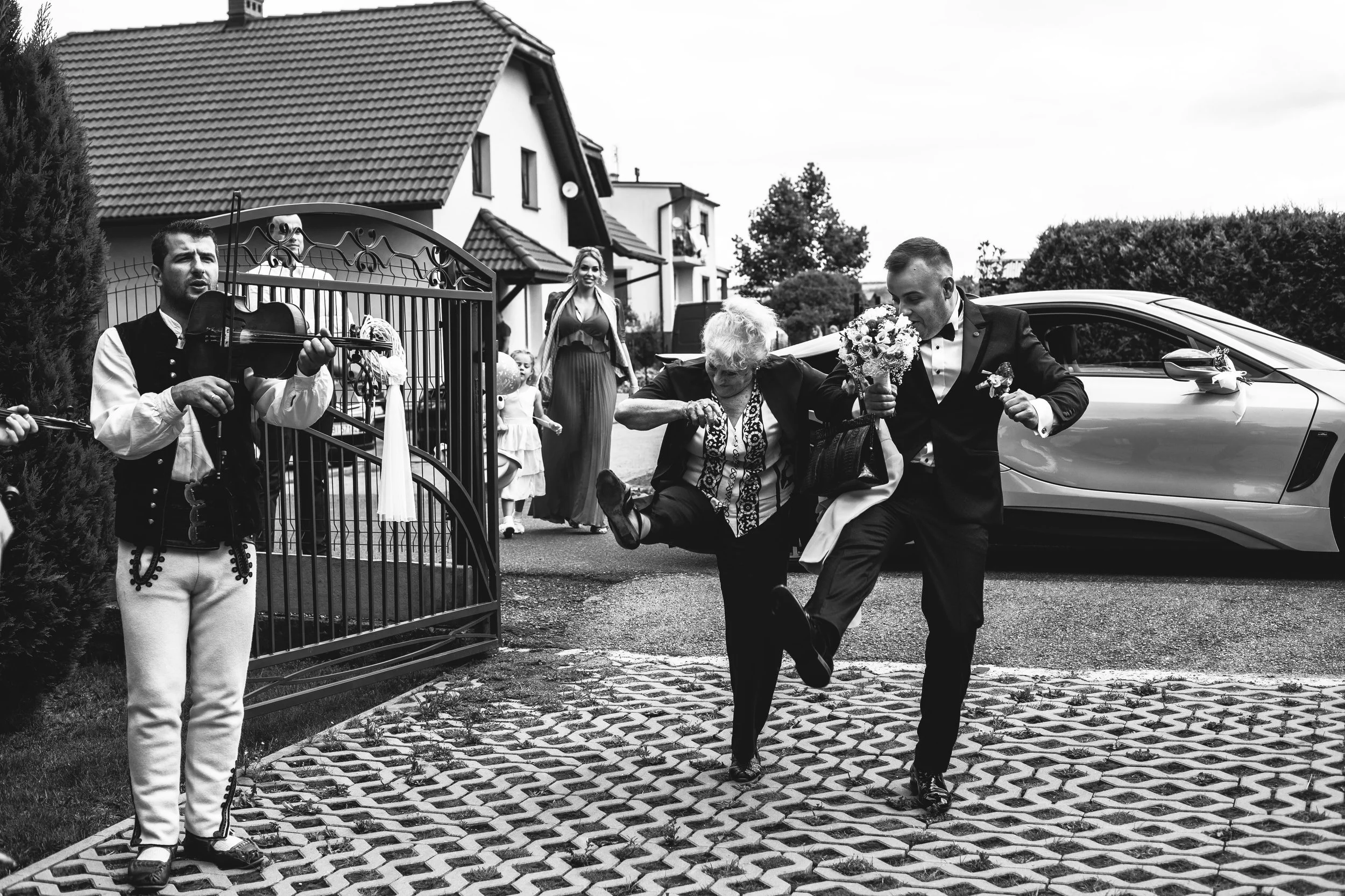 A wedding scene in black and white. A man in a tuxedo is holding a bouquet, stepping over an elderly woman who is standing on the ground. The woman is holding hands with the man. In the background, a violinist dressed in traditional attire is playing