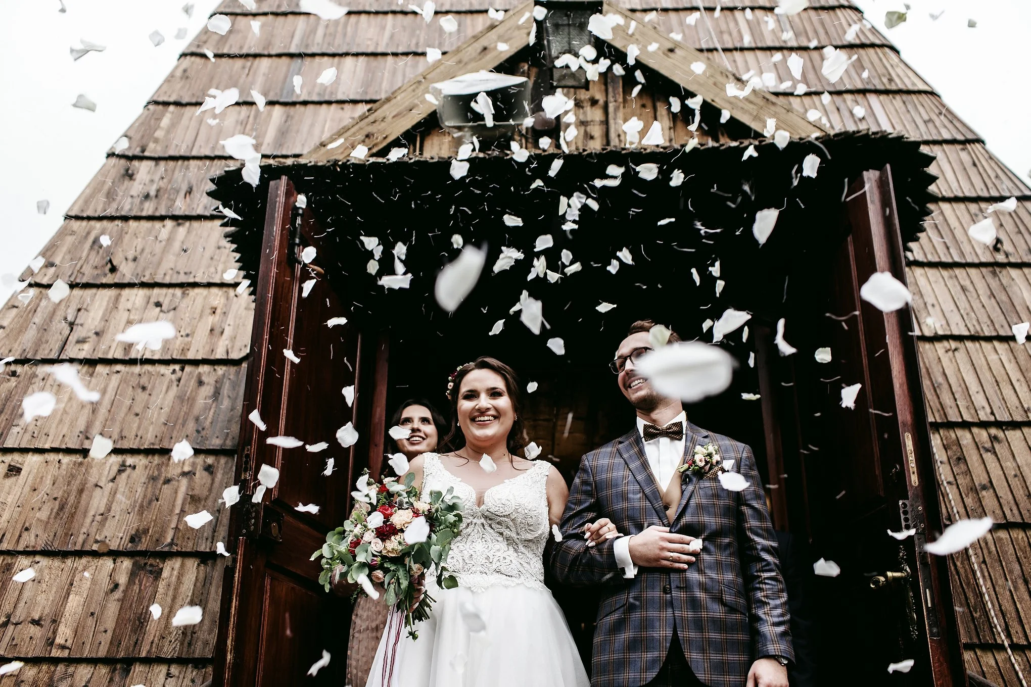 A bride and groom smiling under a shower of white flower petals during their wedding celebration outside a rustic wooden building.