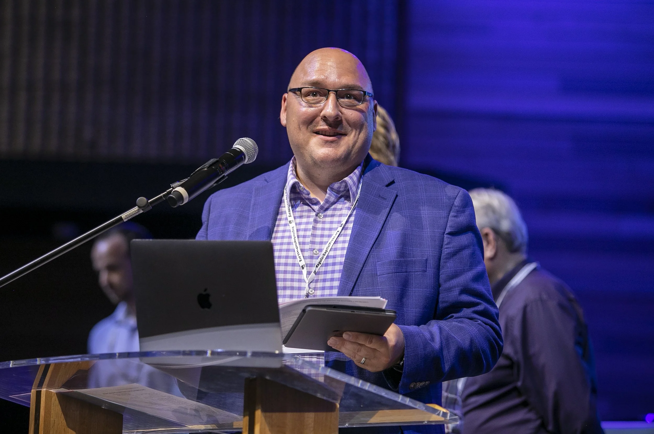 A man wearing glasses, a dark suit, and a colorful floral tie, smiling next to a woman with short hair, wearing a black polka dot blouse with a tie detail, both standing indoors in front of wooden panels.