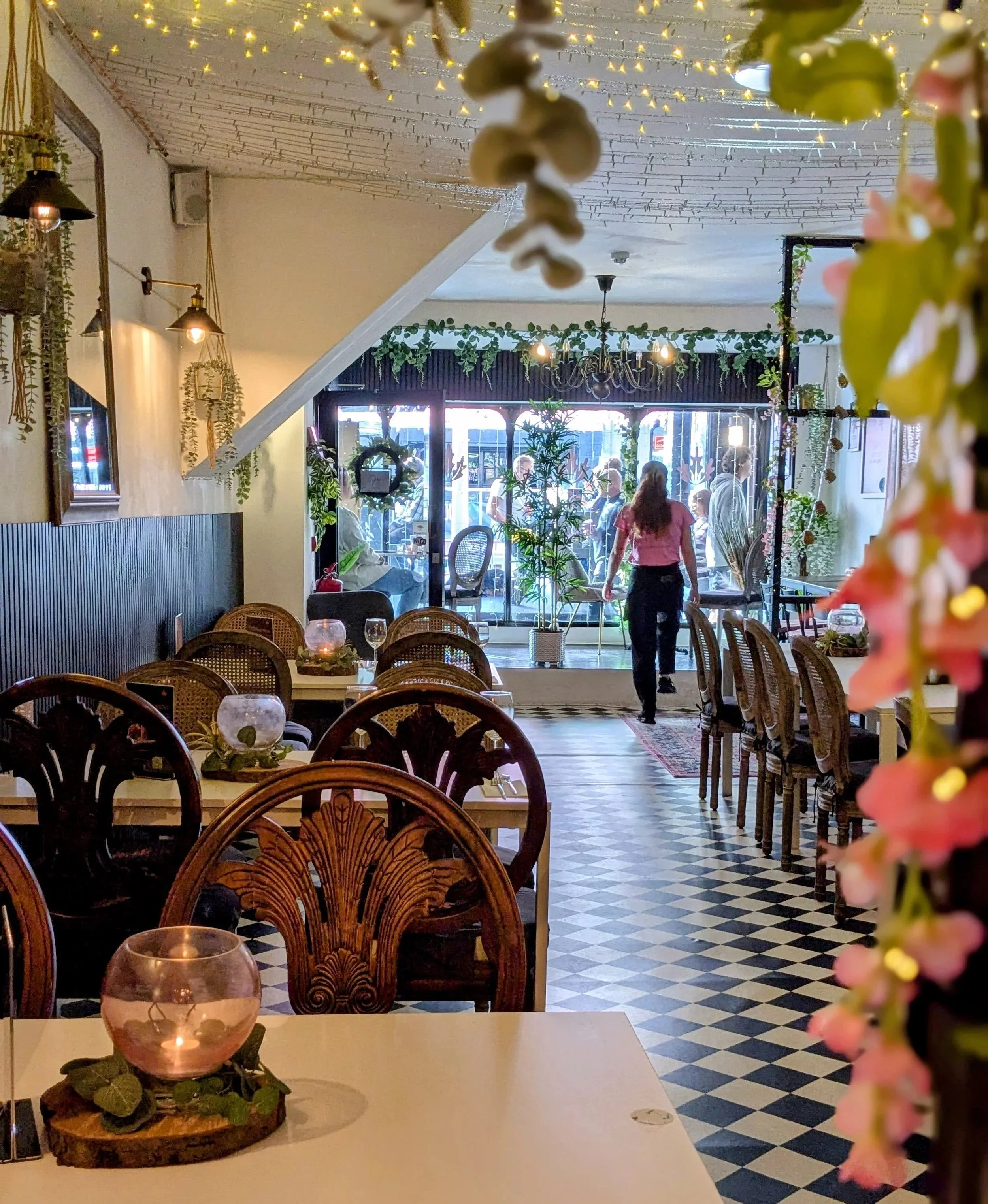 Interior of a cozy restaurant with wooden chairs, tables set with glass candle holders, and a checkered black and white floor. Decor includes hanging plants, framed pictures, and string lights. Behind the glass door, people can be seen outside.