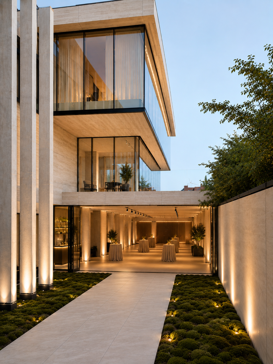 Modern multi-story building with large glass windows, illuminated outdoor corridor with potted plants, tables with tablecloths, and ambient lighting, at dusk.
