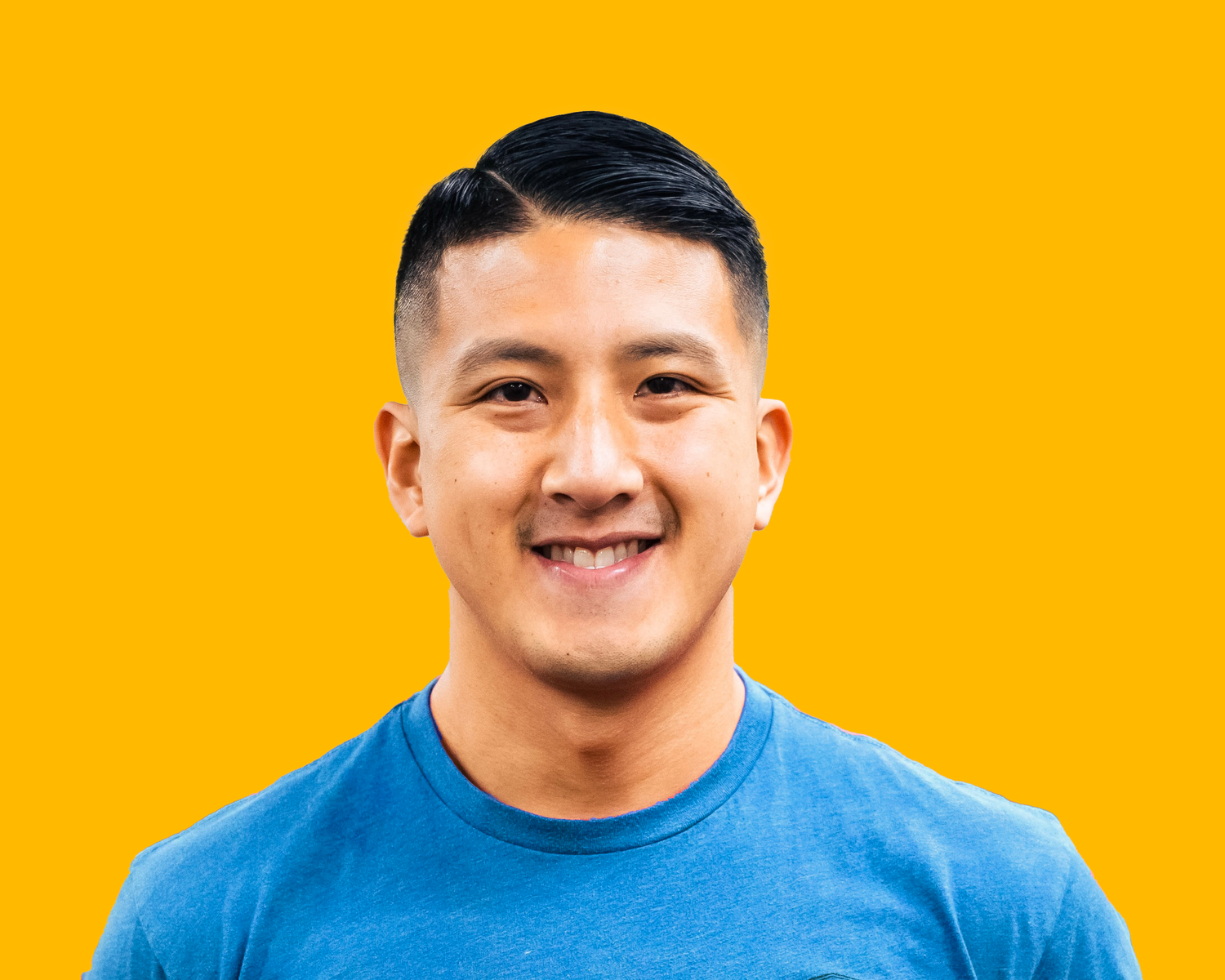Close-up of a smiling young Asian man with short black hair wearing a blue t-shirt against a bright yellow background.