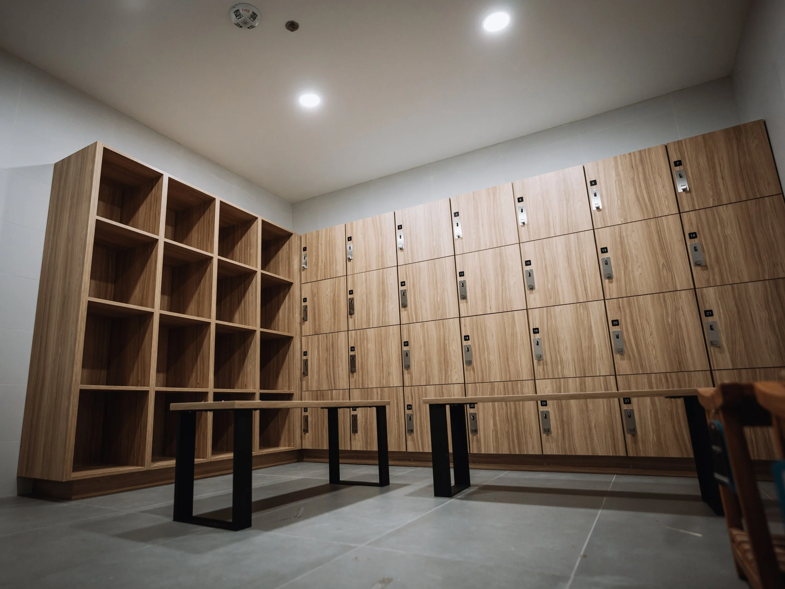 Interior view of a locker room with wooden lockers, some with combination locks, and a table with black legs. The ceiling has bright lighting fixtures.