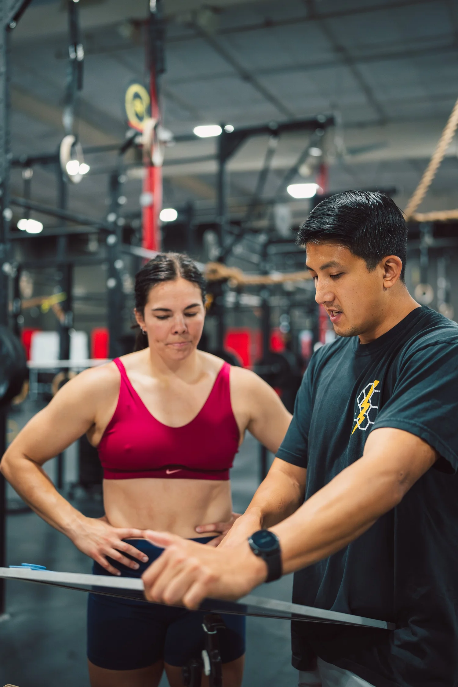A female athlete in a pink sports bra and shorts stands with her hands on her hips, talking to a male trainer or coach in a gym. The man is showing her something on his watch or device. The gym has fitness equipment and ropes in the background.