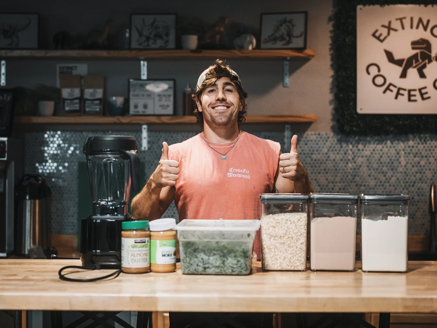 A smiling man with a cap and pink T-shirt giving two thumbs up behind a counter with various food containers and supplements in a coffee shop or cafe.