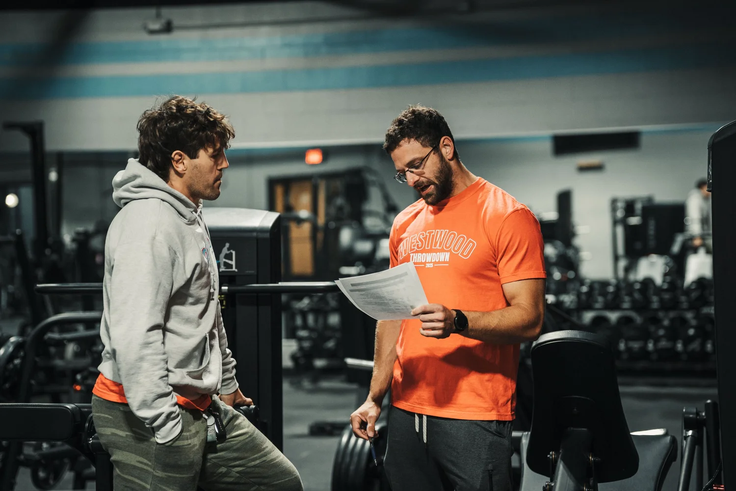 Two men in a gym, one wearing a gray hoodie and camouflage pants, the other in an orange T-shirt with 'Westwood Throwdown' printed on it, reviewing a paper together.