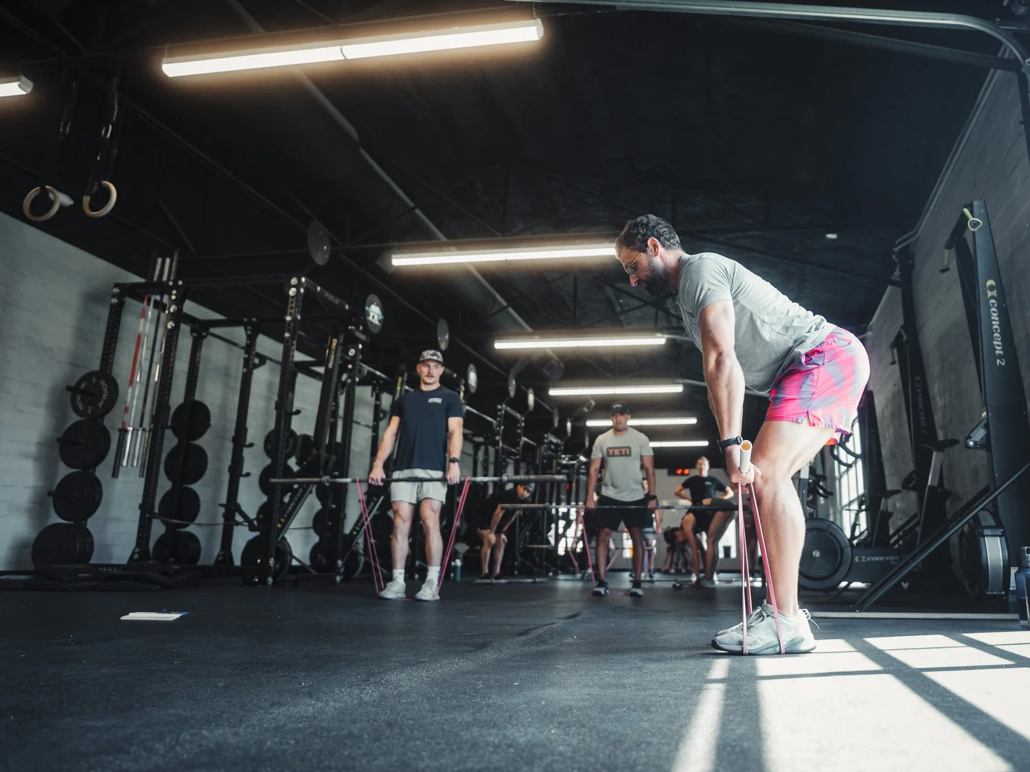 People working out at a gym, performing resistance band exercises on the floor with gym equipment and weight racks in the background.