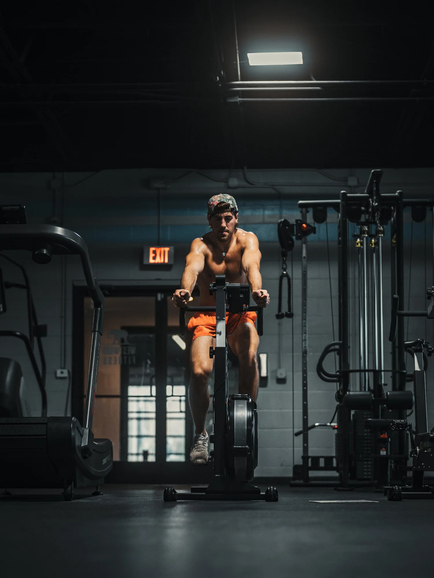 A shirtless man wearing a baseball cap and orange shorts using a rowing machine in a dark gym, with gym equipment and a lit exit sign in the background.