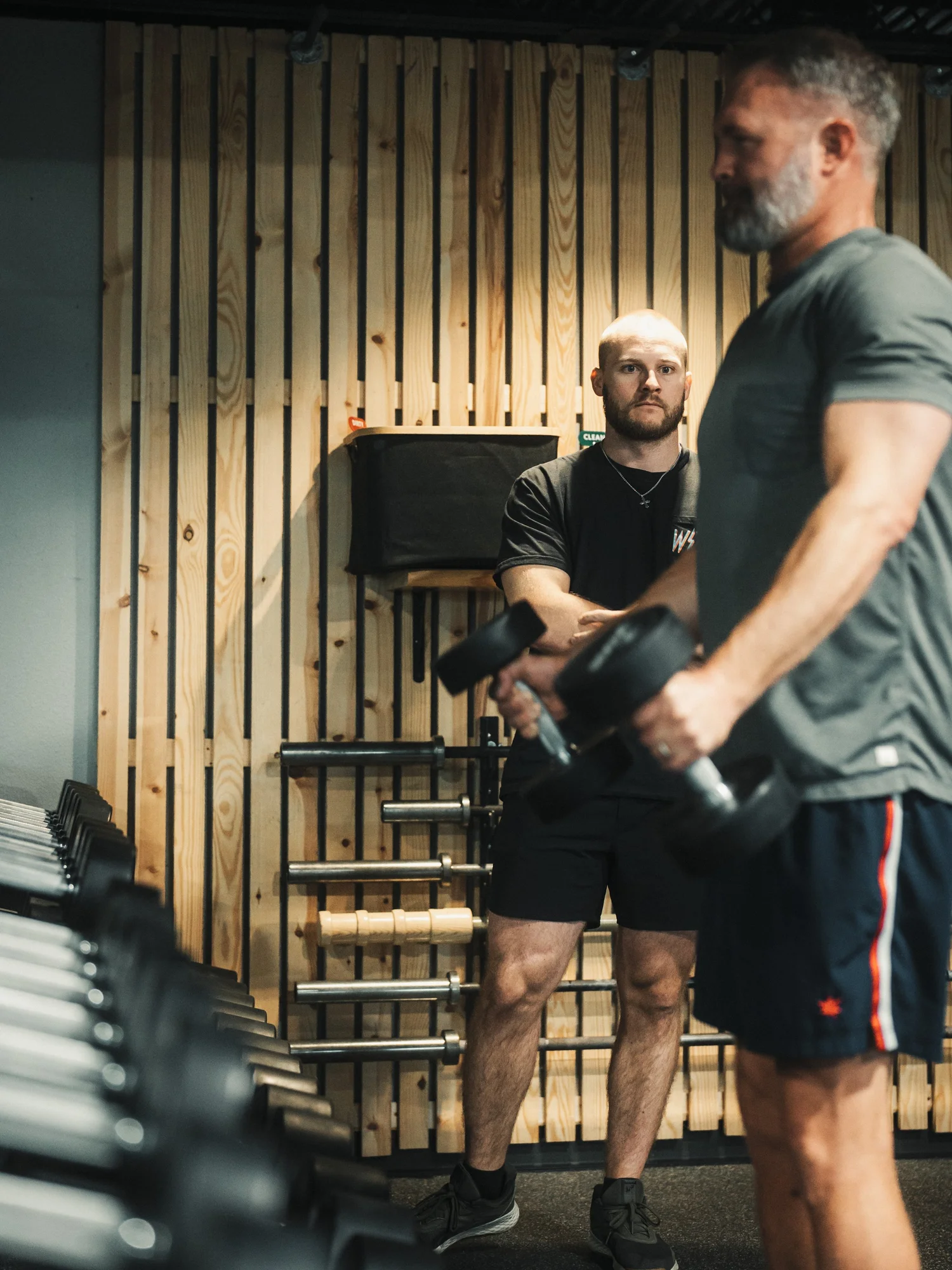Two men in a gym with wooden panel walls, one in the background and one in the foreground. The man in the background has a beard and is looking at the other man, who is holding a weight plate.