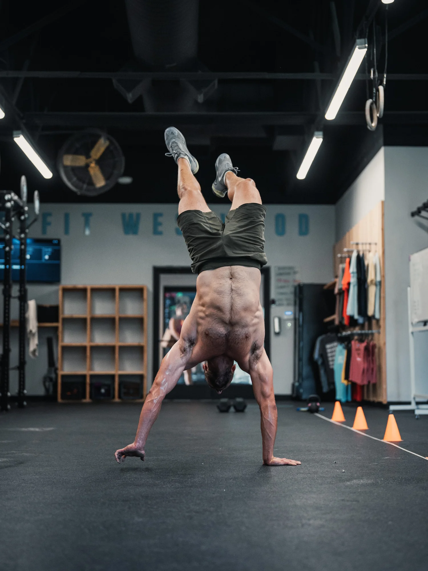 A man doing a handstand in a gym with black walls, gym equipment, and orange cones on the floor.