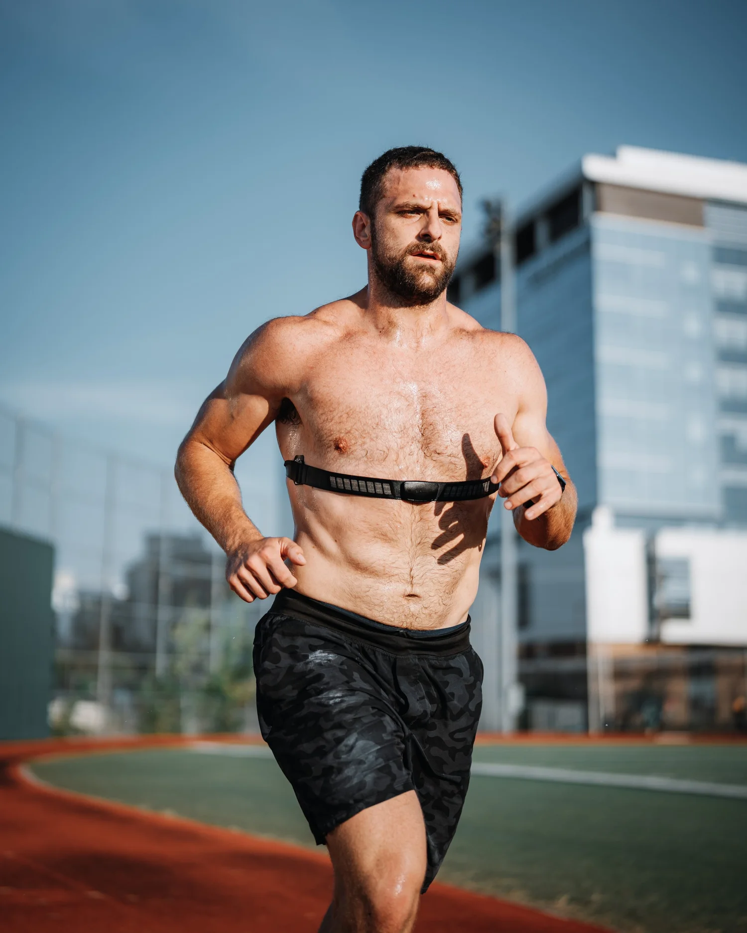 A man running outdoors on a track, wearing black shorts and a running harness, with modern buildings in the background and a blue sky.
