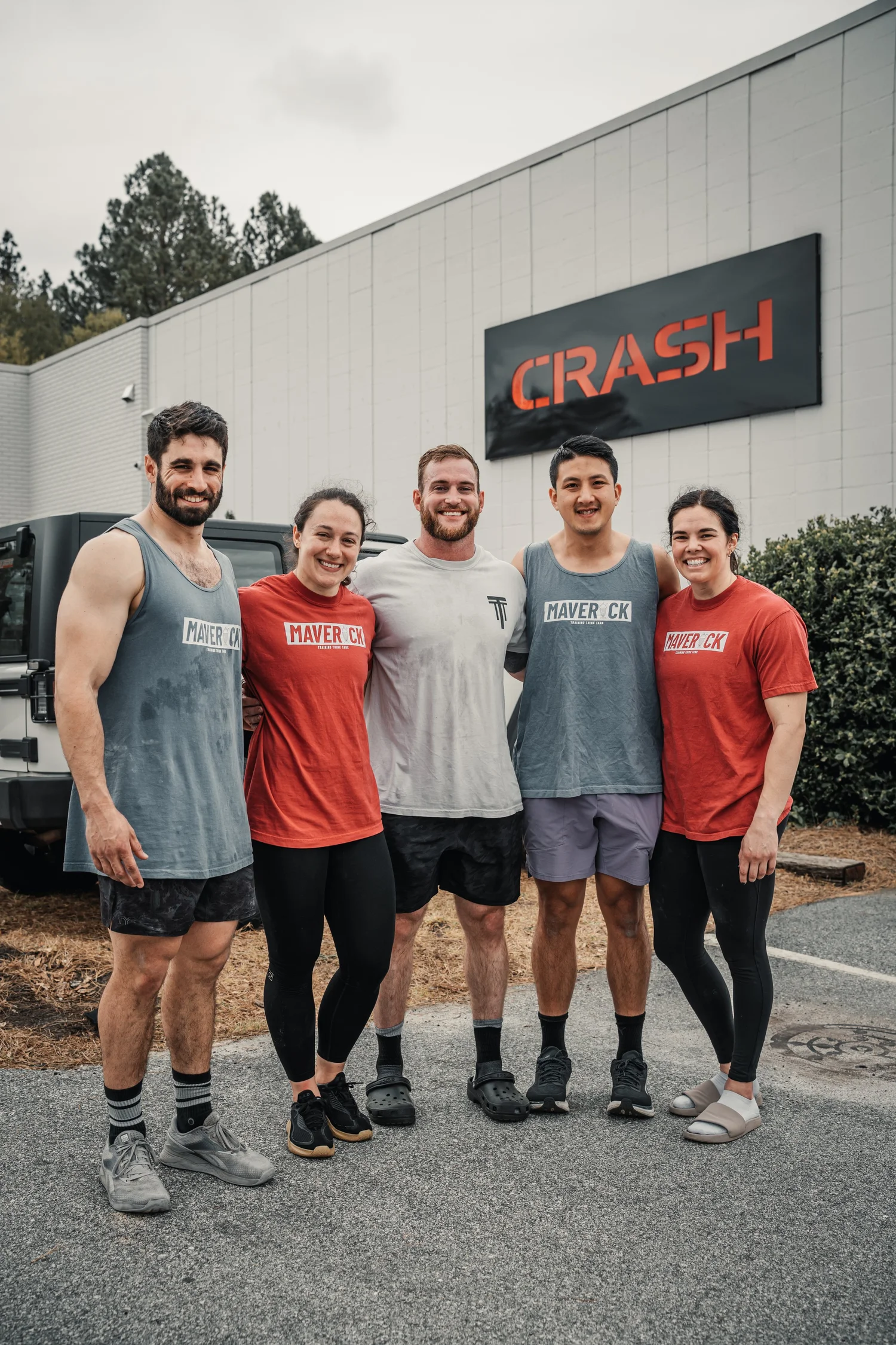 Group of five fitness enthusiasts standing outside near a white building with a large sign that reads 'CRASH'. They are dressed in workout clothes, some wearing tank tops and leggings, smiling at the camera.