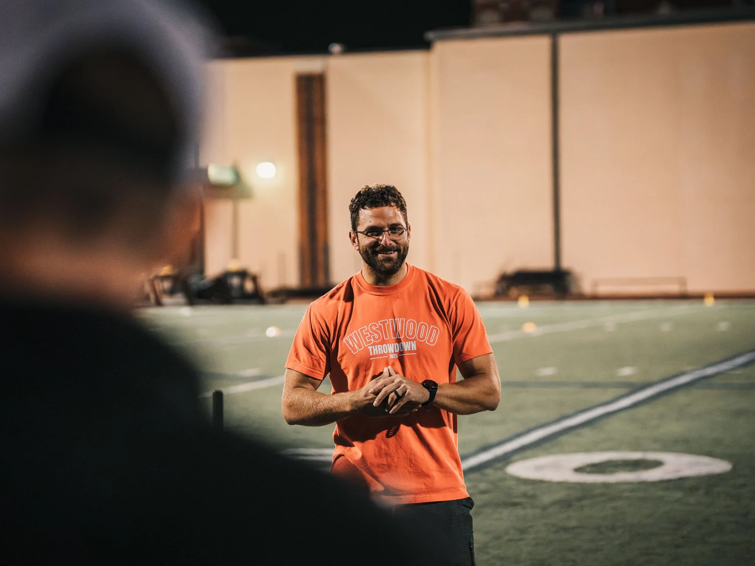 A man in an orange shirt with 'Westwood Throwdown 2023' written on it, smiling and standing outdoors at night on a sports field, with a blurred figure in the foreground and a large beige building in the background.