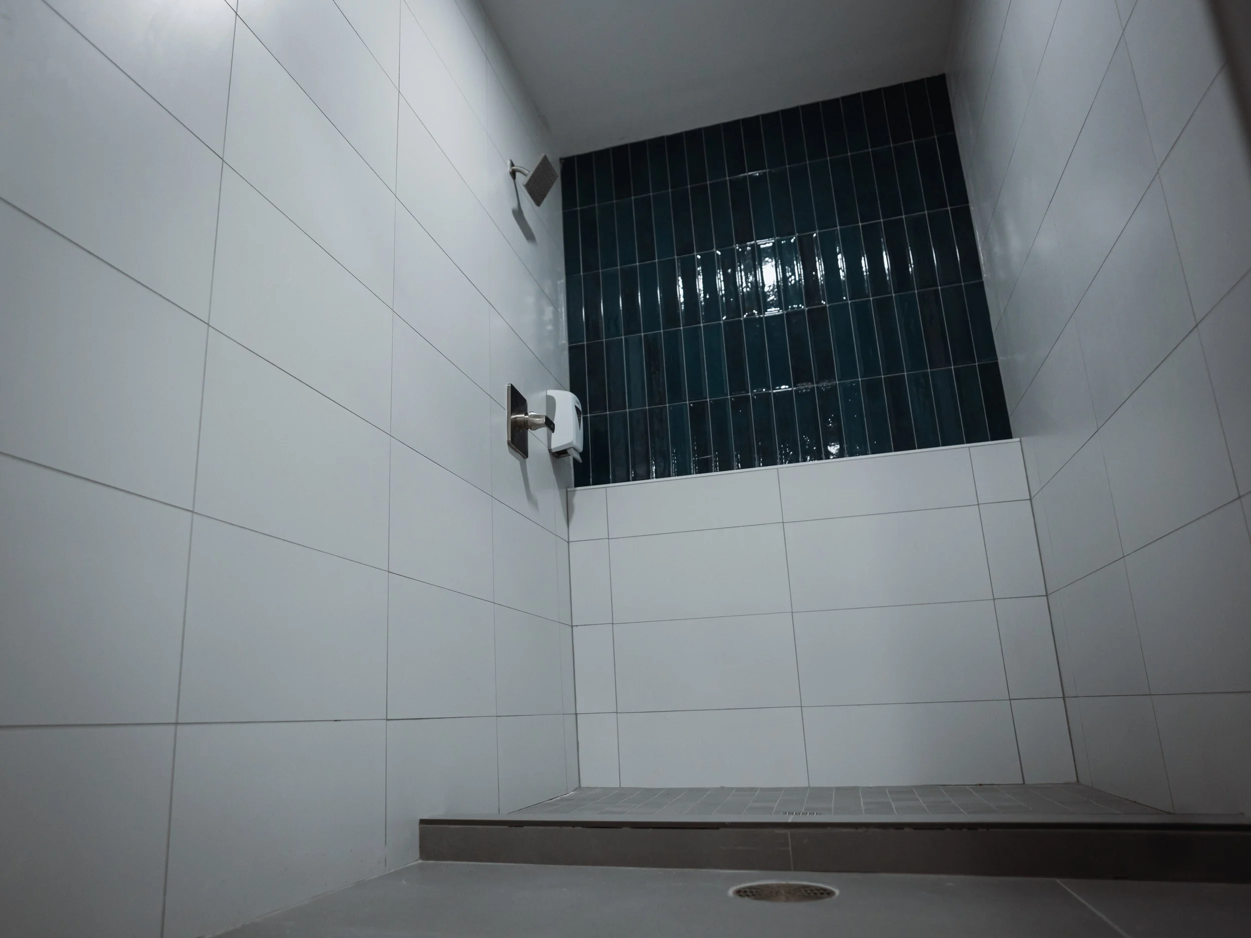 Empty shower stall with white and dark blue tile walls, a showerhead, a soap dispenser, a drain on the floor, and a dark grey ledge.