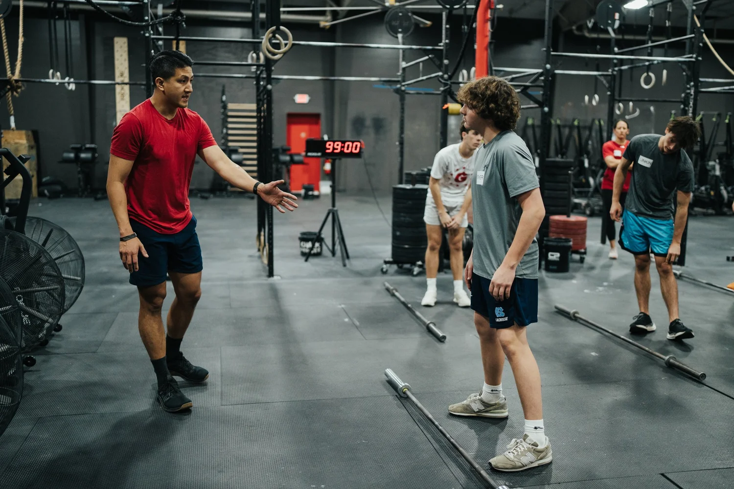 Two athletes are having a conversation in a gym during a workout. One is wearing a red shirt and black shorts, and the other is in a gray shirt and black shorts. The gym has weightlifting equipment and a digital timer showing 99:00 in the background.