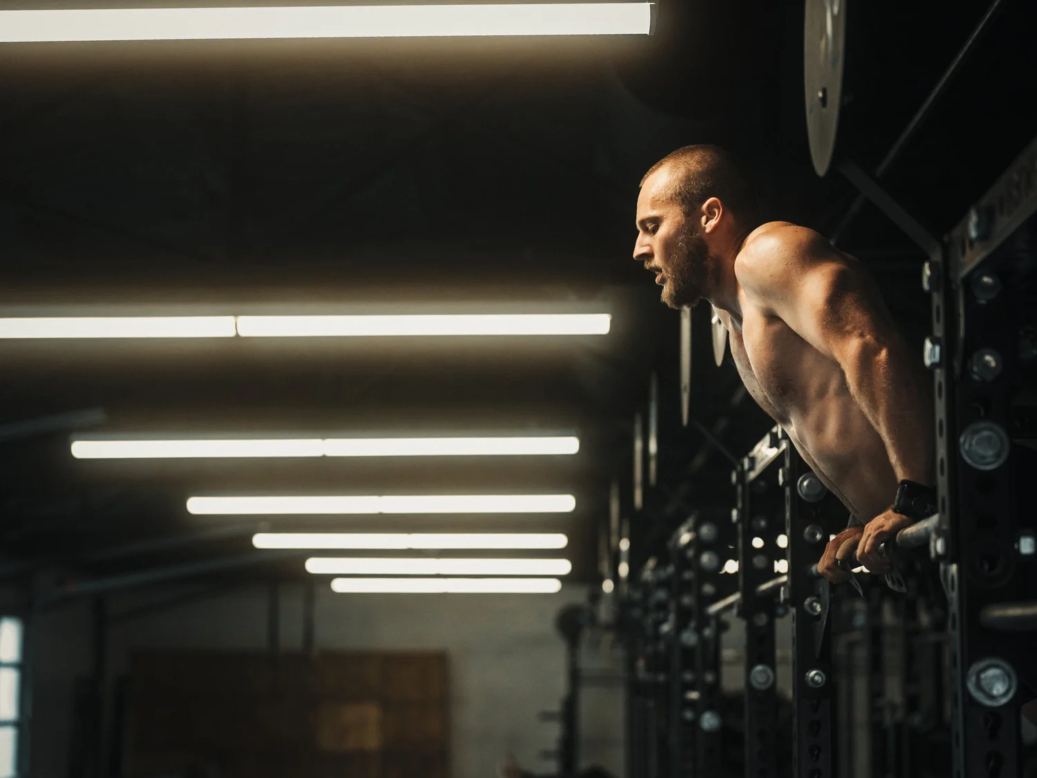 A man with a shaved head and beard doing push-ups in a dimly lit room between metal racks with fluorescent lighting.