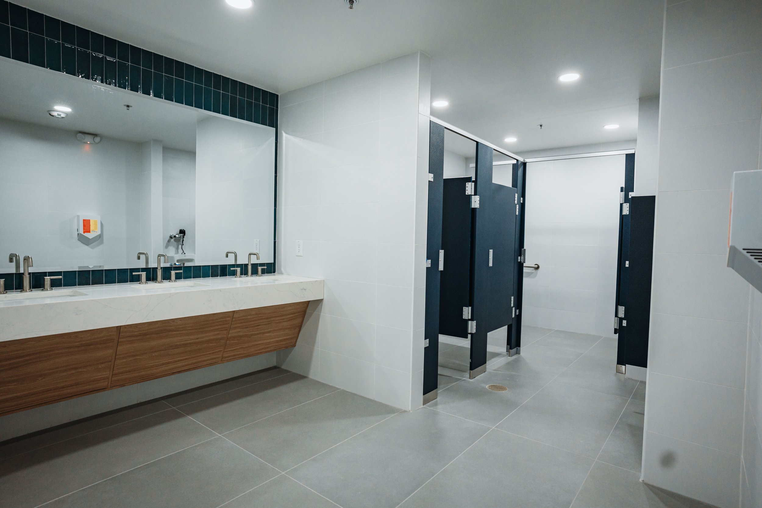 Public restroom with multiple stalls, a row of sinks, and white tiled walls.
