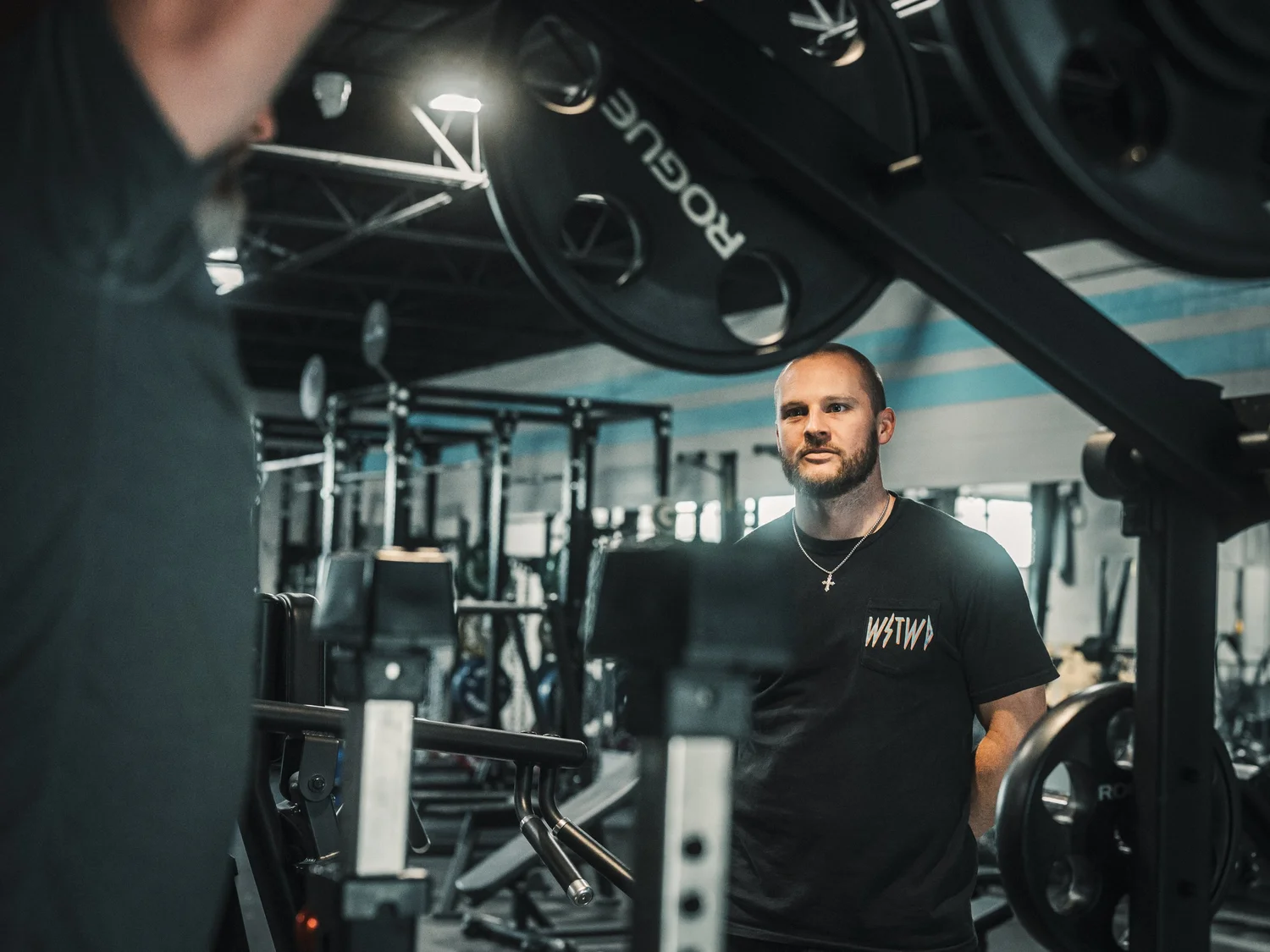 A man with a beard and cross necklace at the gym, standing near weightlifting equipment, with large machinery in the background.