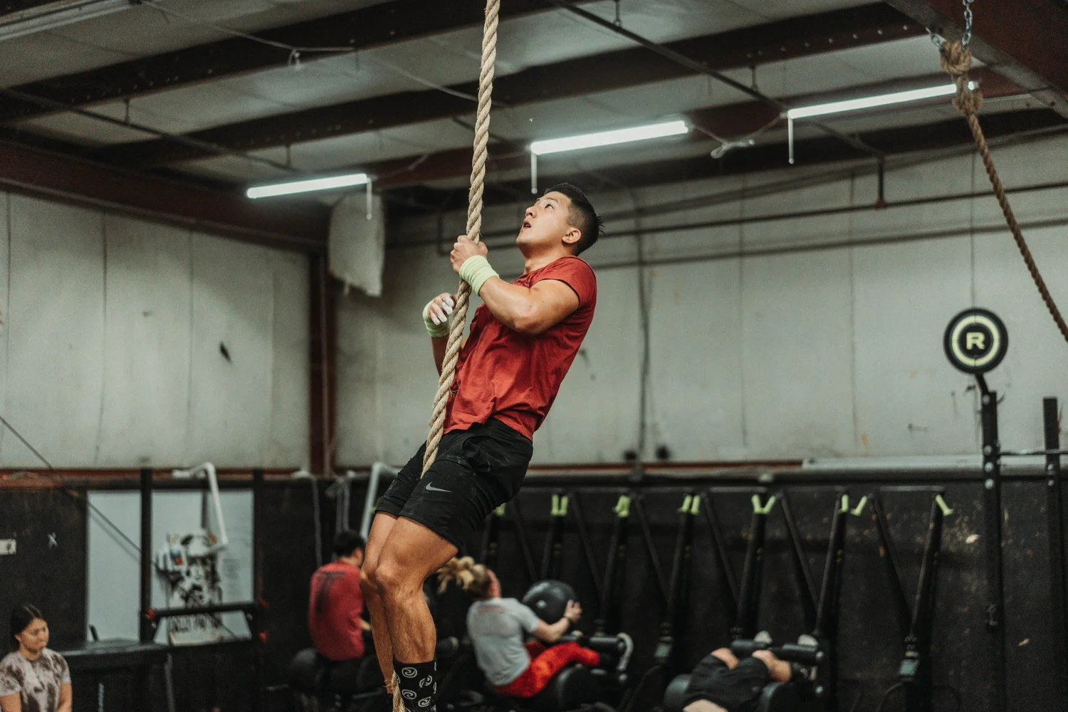 A man in a red shirt and black shorts climbing a thick rope inside a gym or fitness center, with several individuals and gym equipment visible in the background.