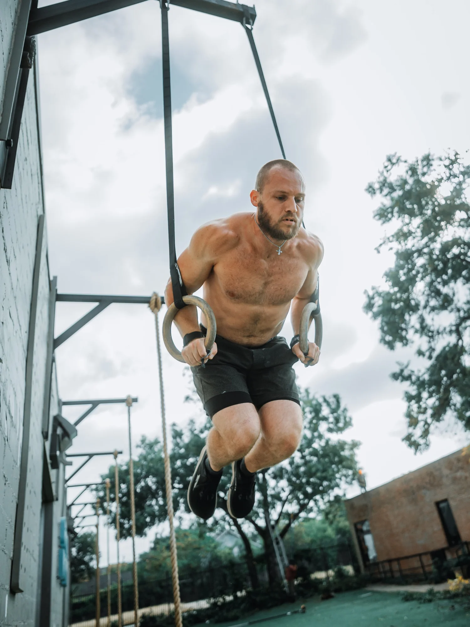 A shirtless man with a beard and a cross necklace is doing a workout on gymnastic rings outdoors, looking focused under a cloudy sky.
