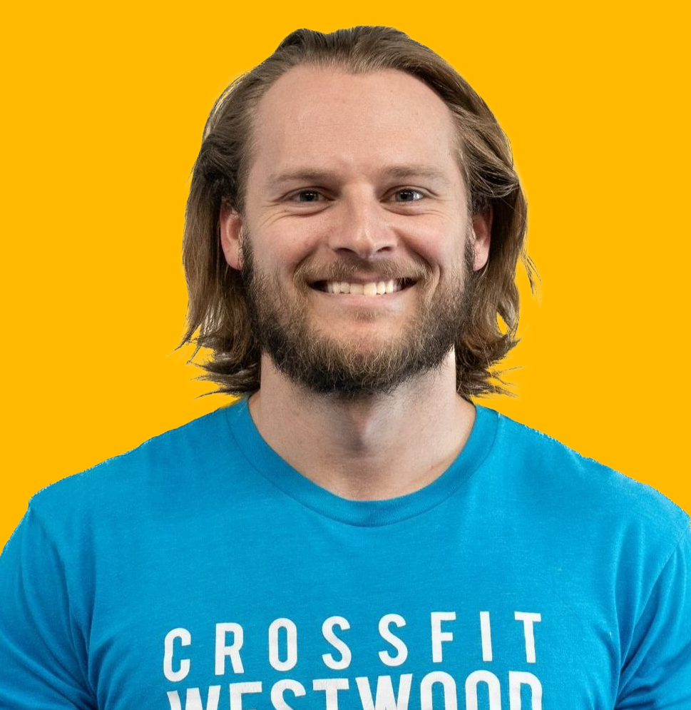 A man with long hair and a beard smiling, wearing a blue CrossFit Westwood t-shirt against a yellow background.