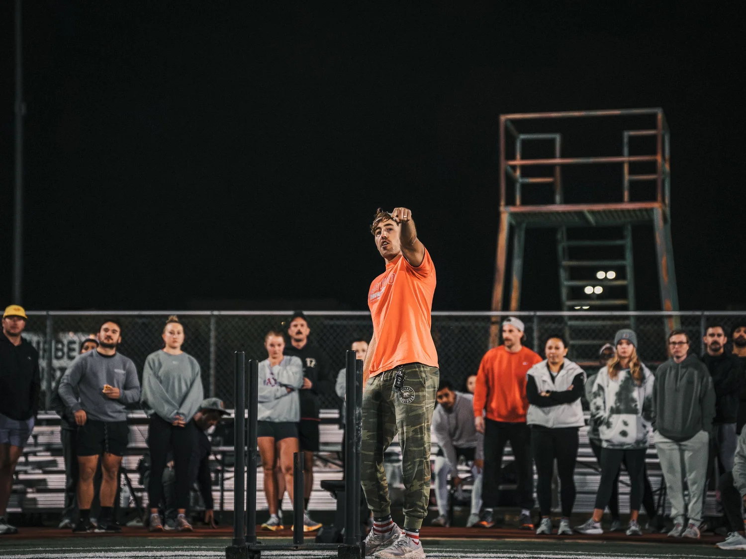 A man in an orange shirt and camouflage pants is pointing at the camera while standing in front of a group of spectators at night. The crowd is watching him on a sports field, with a metal structure and bright lights in the background.
