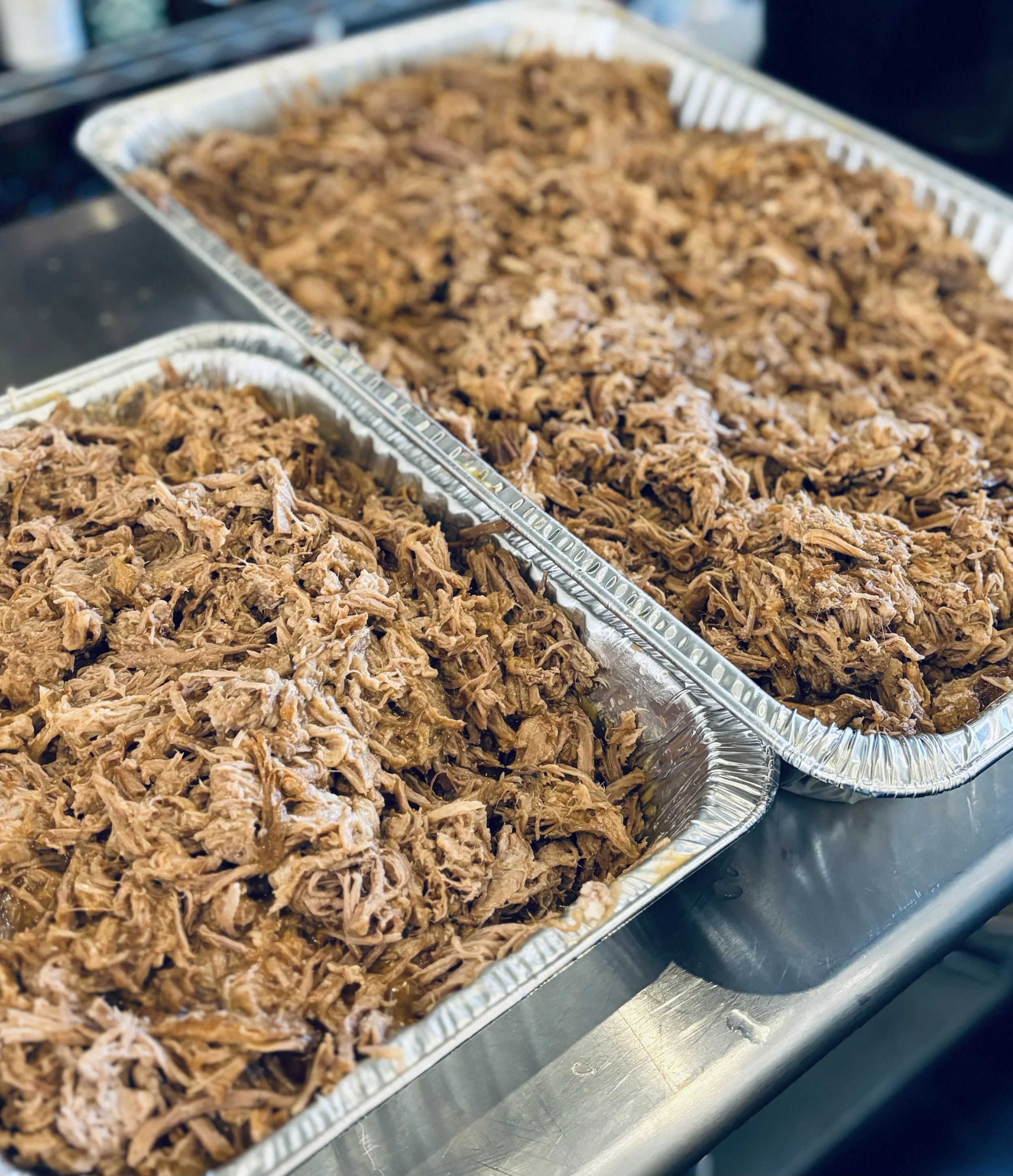 Two aluminum trays filled with shredded cooked meat, likely pulled pork or beef, placed on a stainless steel countertop.