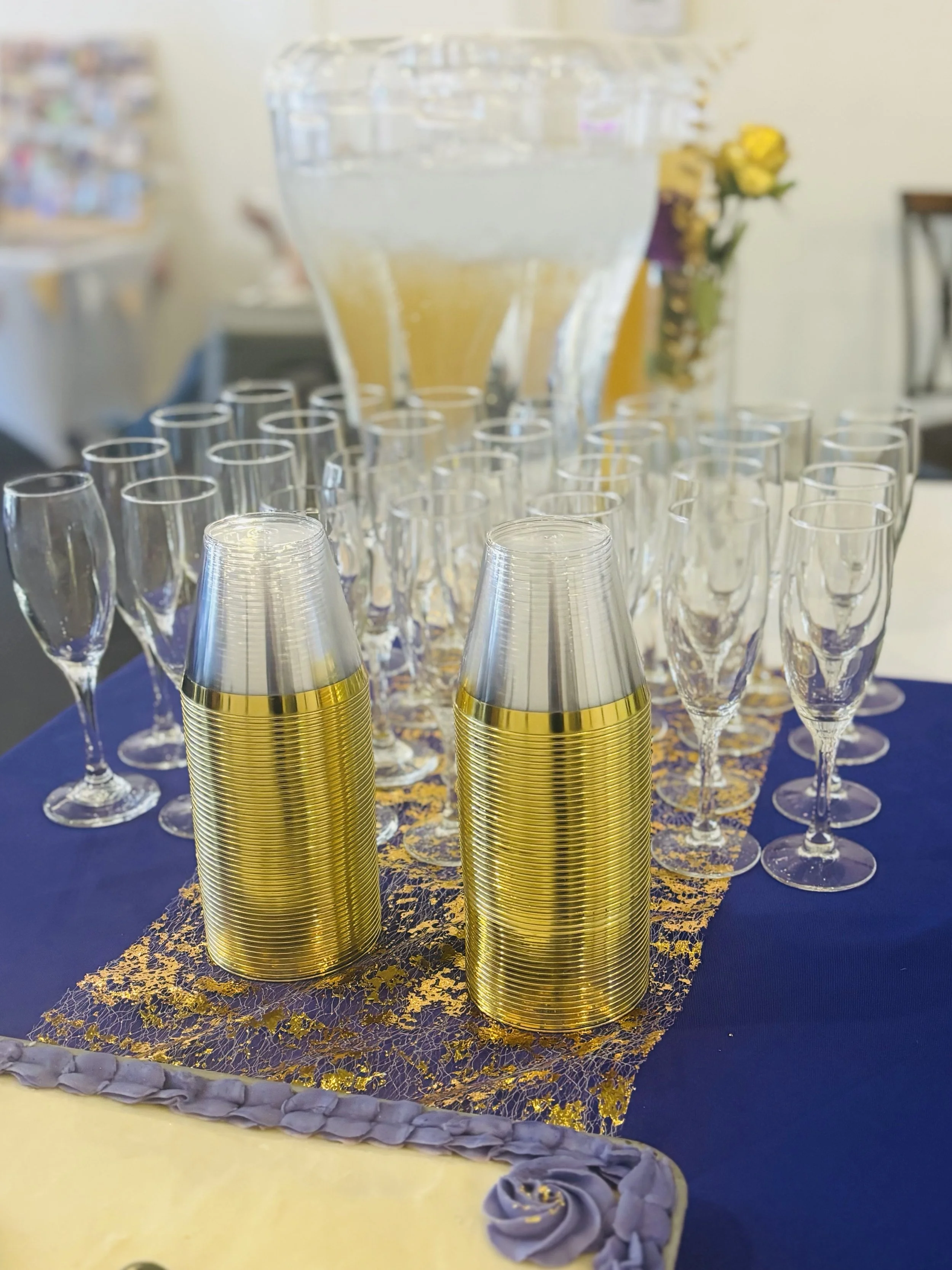 A table with a blue tablecloth, gold and clear disposable cups, and a large glass beverage dispenser filled with a light-colored liquid, possibly juice or water, in the background.