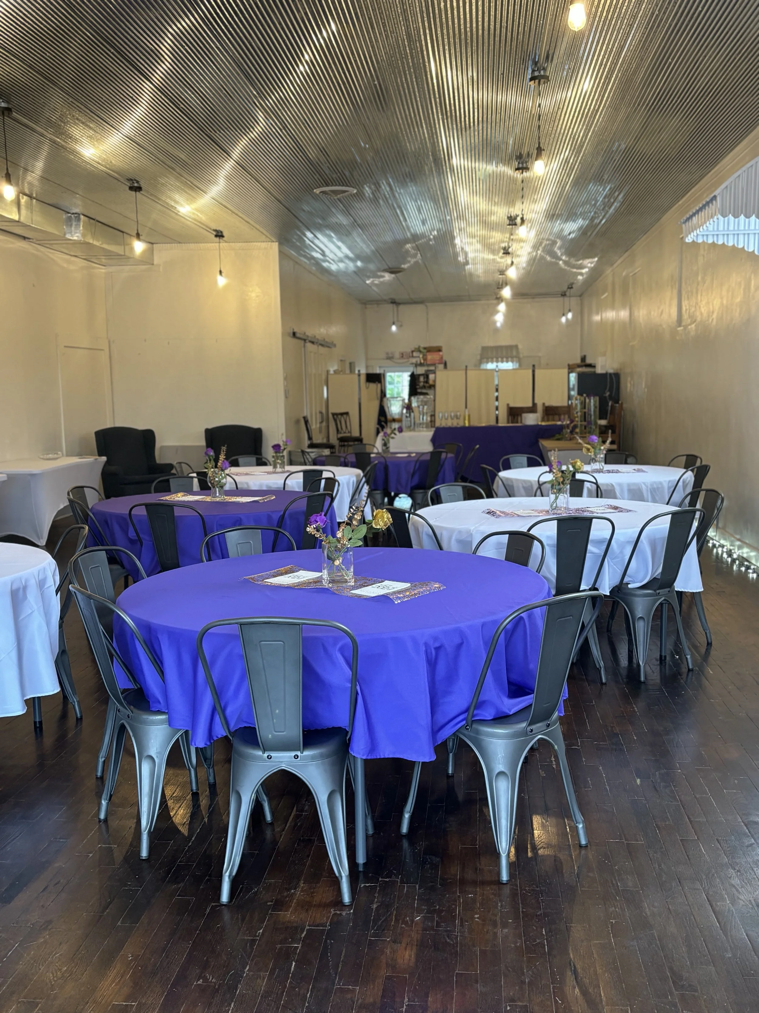 Event space with round tables covered in purple and white tablecloths, each decorated with small flower arrangements, set for a celebration or gathering. The room has a wooden floor and a corrugated metal ceiling with hanging light bulbs, with a small window at the back.