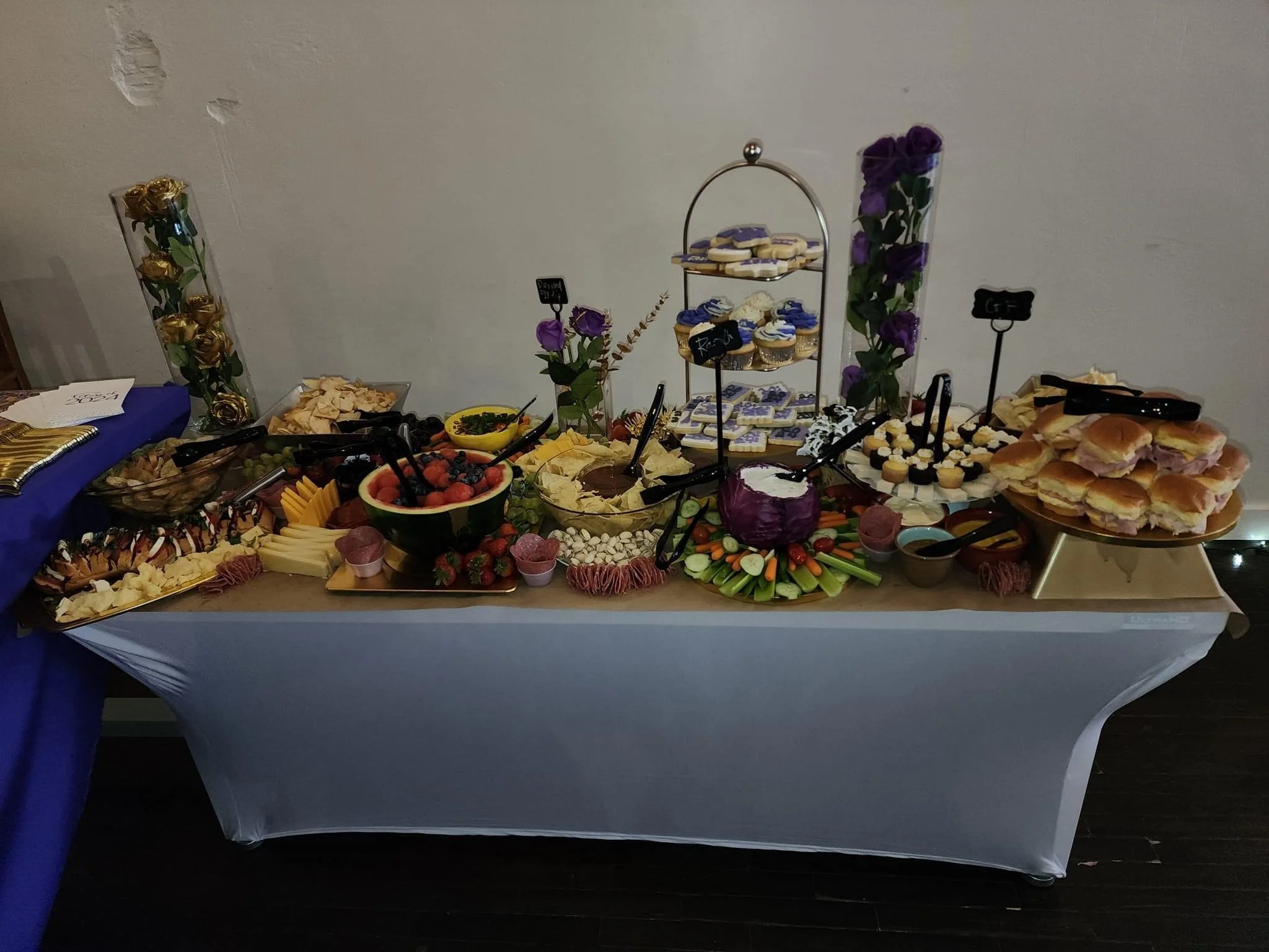 A buffet-style food table with various snacks and desserts, including a bowl of mixed berries, sandwich sliders, vegetables, cheese, chips, cupcakes, cookies, and decorated with purple flowers and gold accents.
